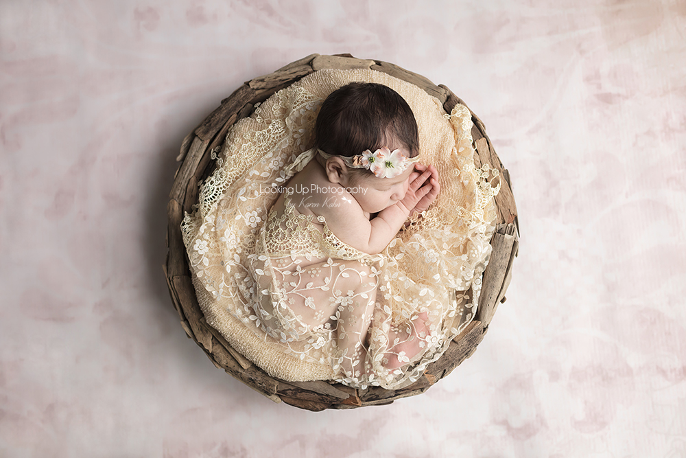 Birds eye view posed newborn in circle wooden basked surrounded by gold lace with adorable hair and flower headband and clasping hands on pink backdrop for baby girl portrait session
