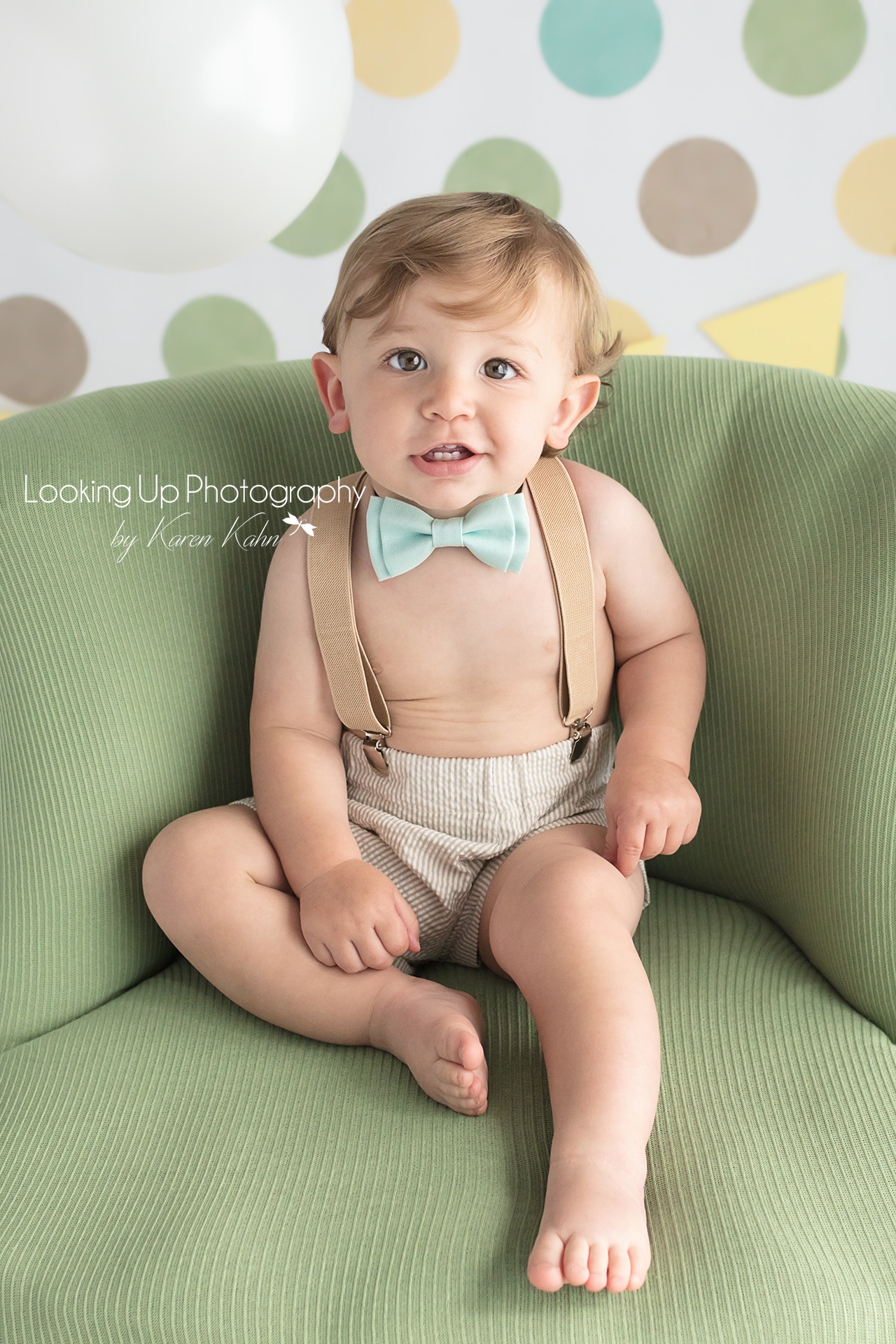 One year old baby boy wearing bow tie and suspenders smiling in snazzy green chair and polka dots for 12 month milestone portrait cake smash session