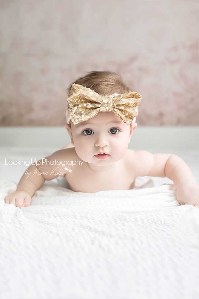 4 month milestone portrait session of baby girl laying on her stomach with gold glitter bow headband and pink backdrop