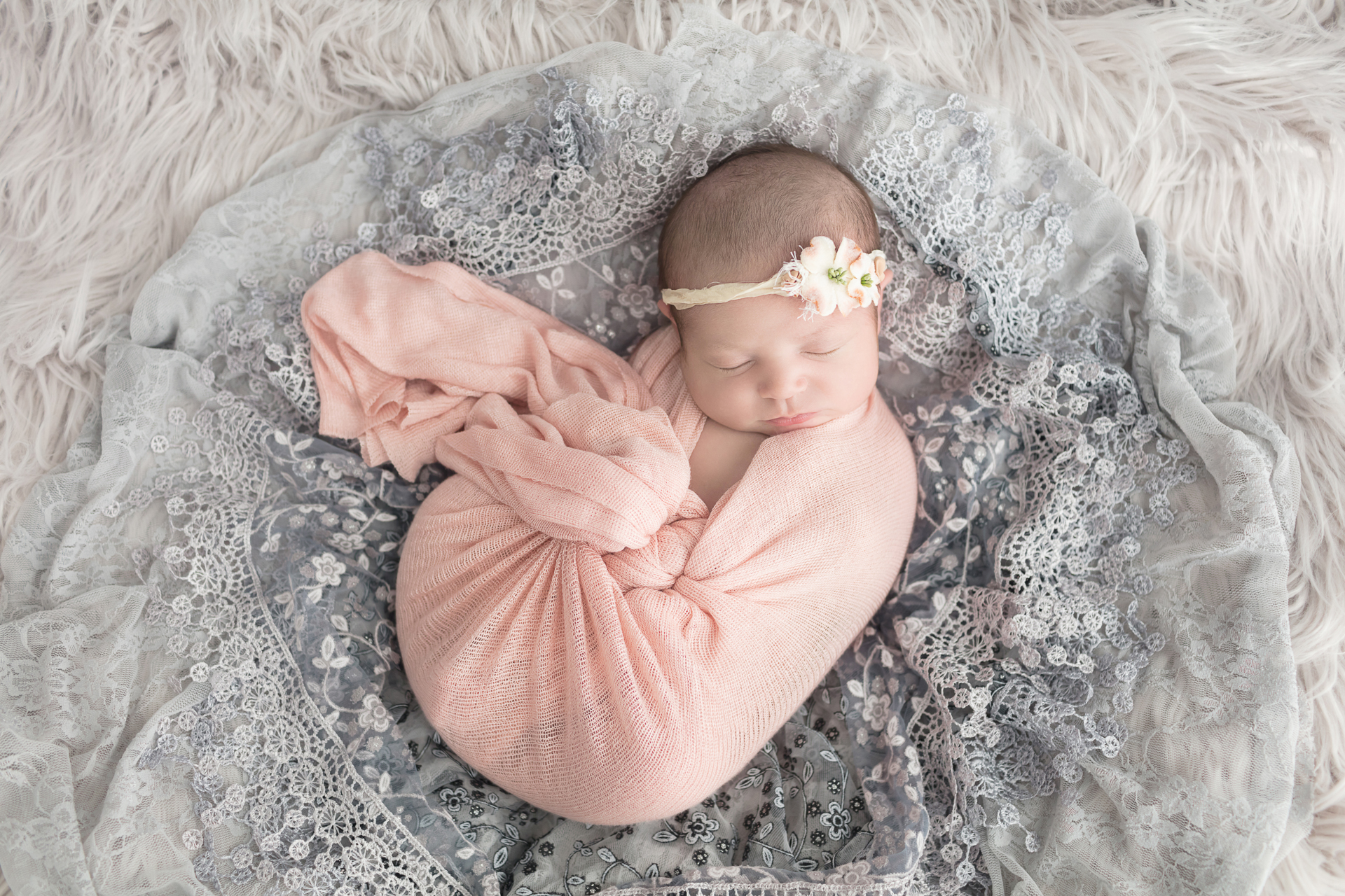 Newborn portrait session of baby girl with peach and white flower headband swaddled in peach wrap surrounded by gray lace and gray fur