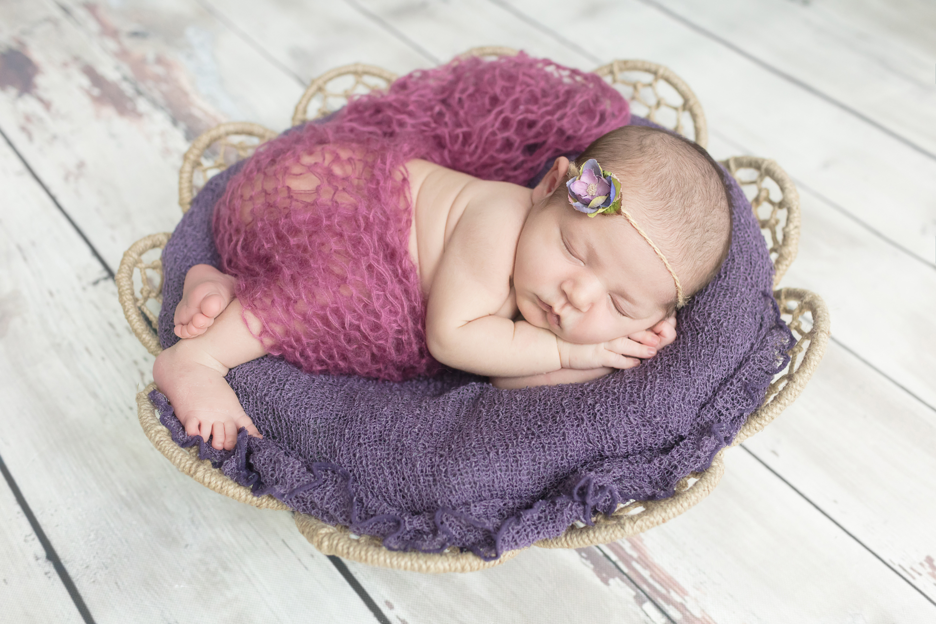 Newborn portrait session of baby girl posed in a flower shaped woven basket and purple and violet wraps, with with purple flower headband on white wooden floor