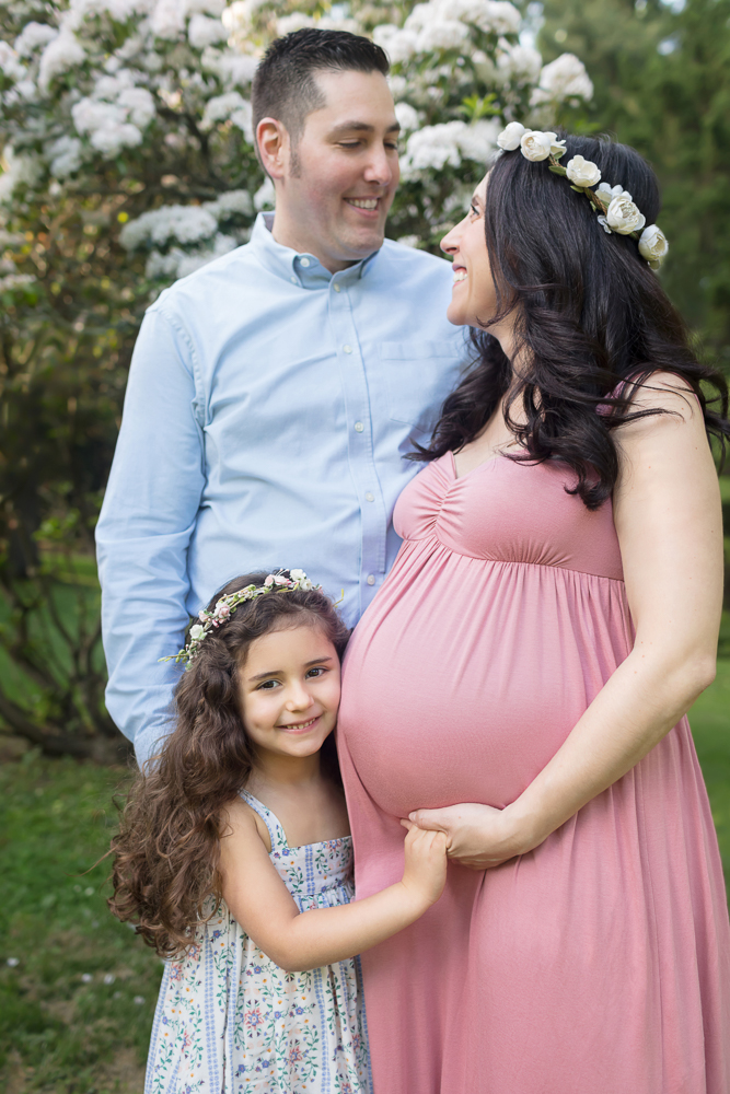 Smiling big-sister-to-be and expecting mom in pink with matching flower wreaths, and dad in front of lovely blooming tree of flowers for posed outdoor maternity session