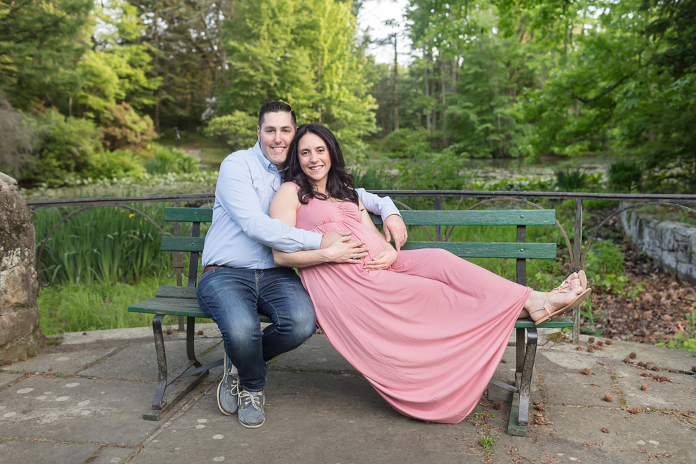 Smiling expecting mom in pink and dad lounging on bench enveloped in gorgeous green spring colors for posed outdoor maternity session