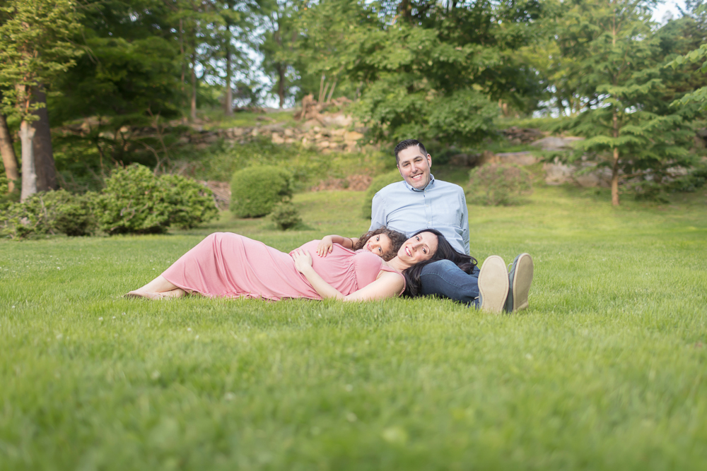 Posed outdoor maternity session with happy expecting mom in pink, big-sister-to-be, and dad lounging on grass in a beautiful field of trees with green spring colors