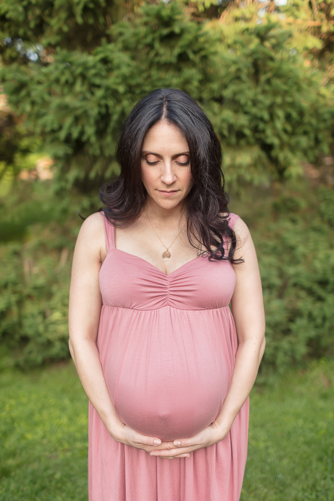 Stunning pregnant mom in pink gown cradling belly surrounded by lovely spring green trees for posed outdoor maternity session