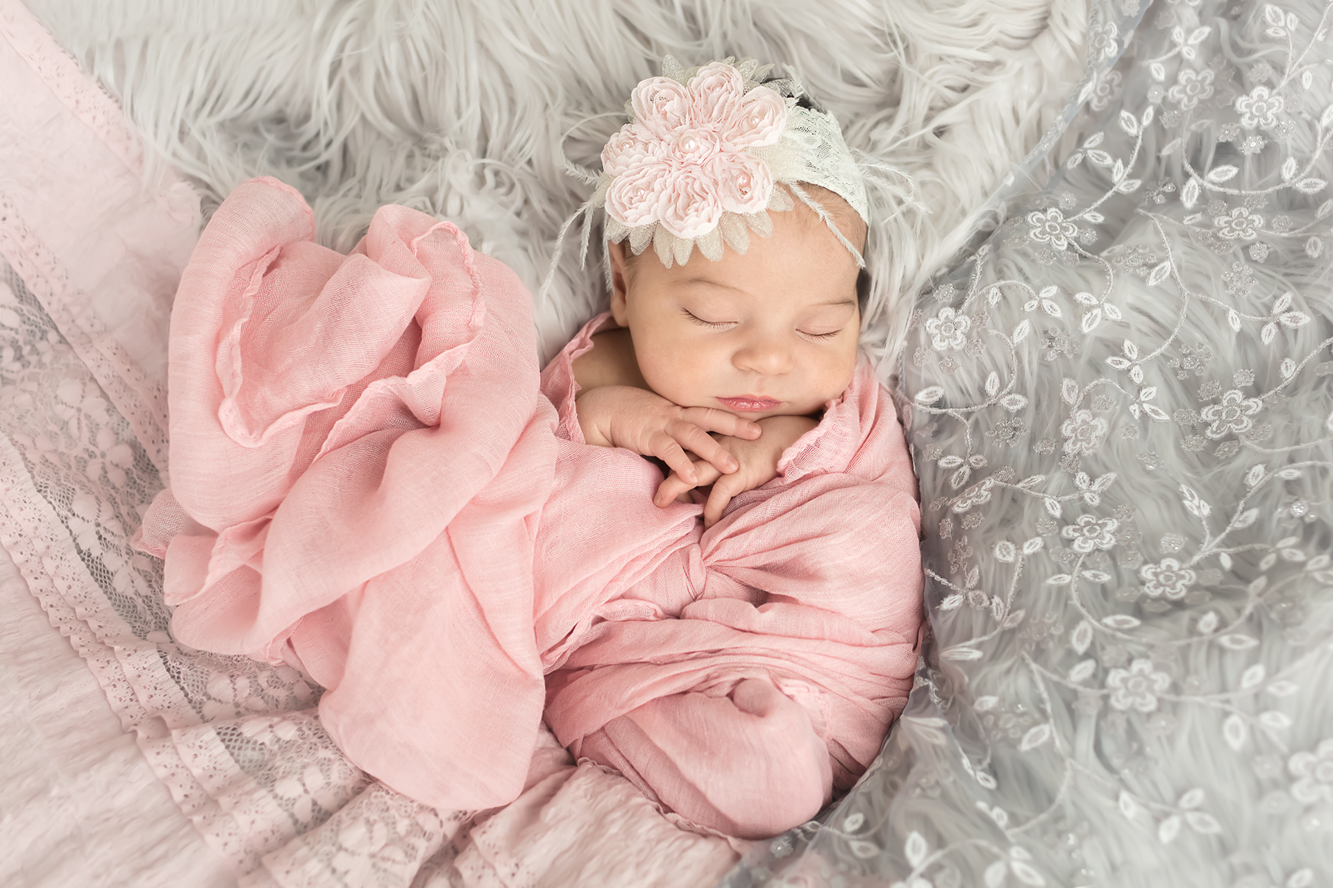 Baby girl swaddled in a pink wrap shaped like a heart and a gorgeous pink and white flower headband peach surrounded by gray and pink lace and gray fur for a Newborn portrait session