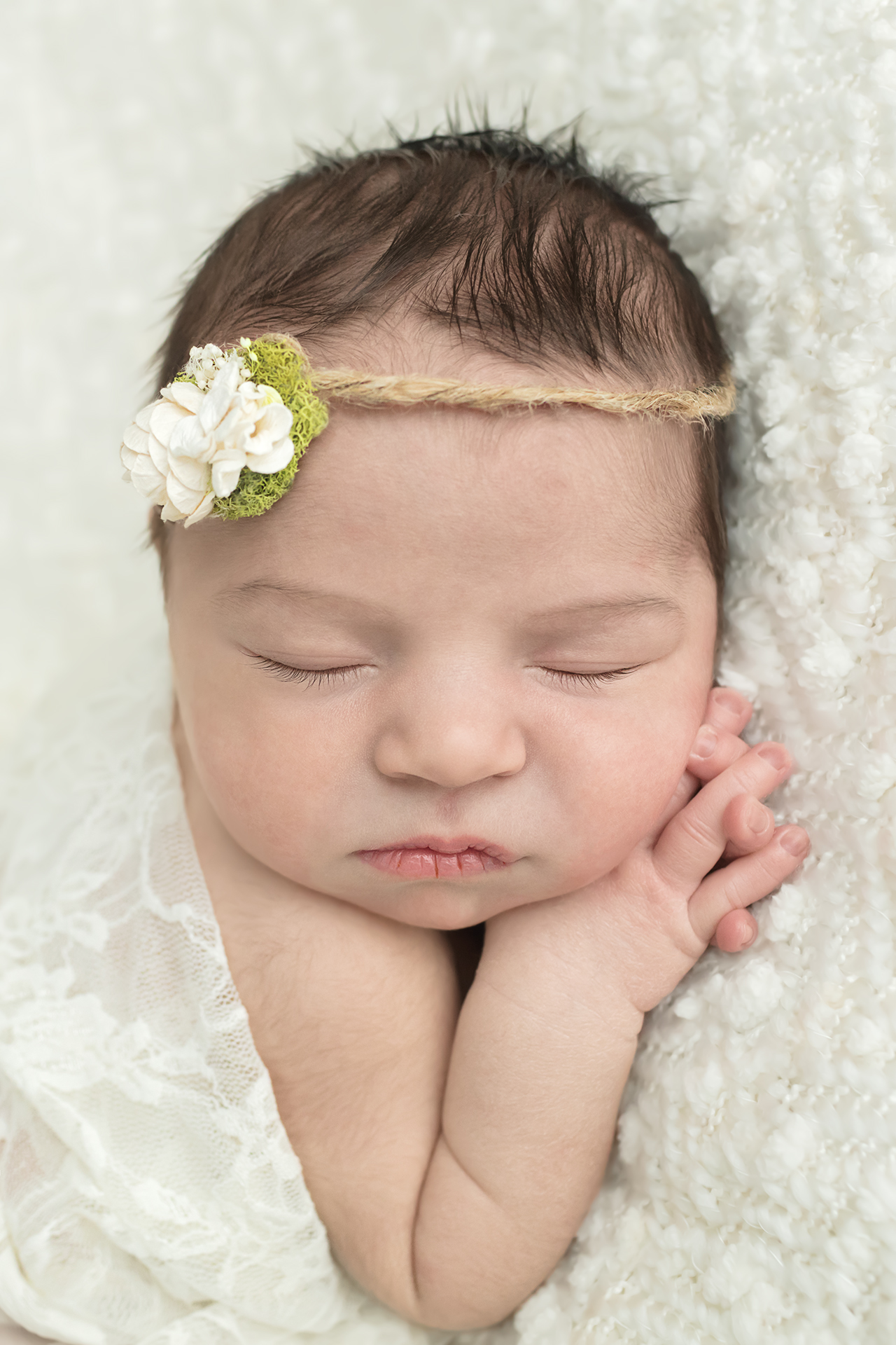 Posed newborn portrait session of baby girl on cream blanket with white flower headband with close up of hand clasped and adorable face