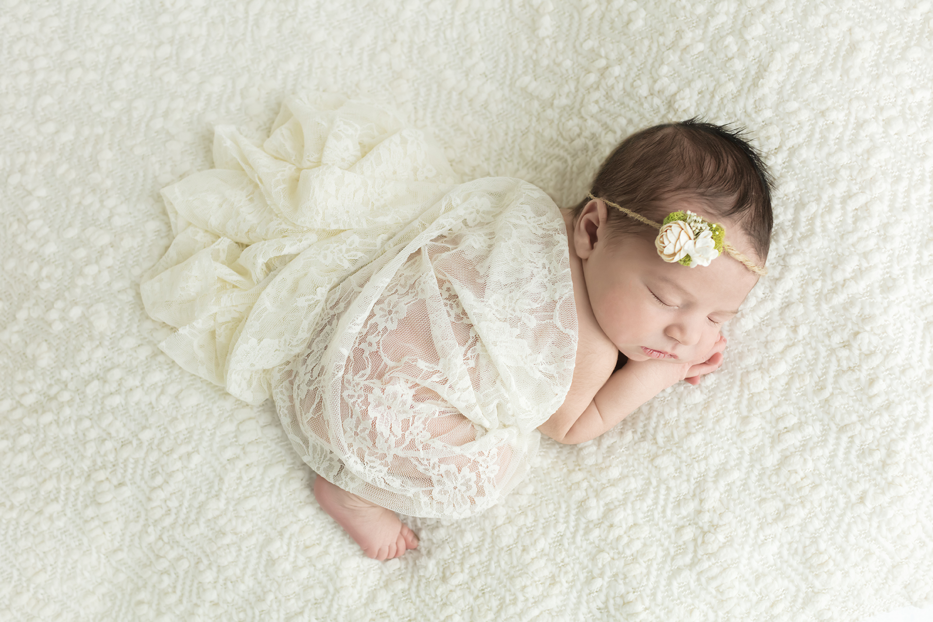 Sleeping baby girl posed wrapped in cream lace on cream blanket with white flower headband for newborn portrait session