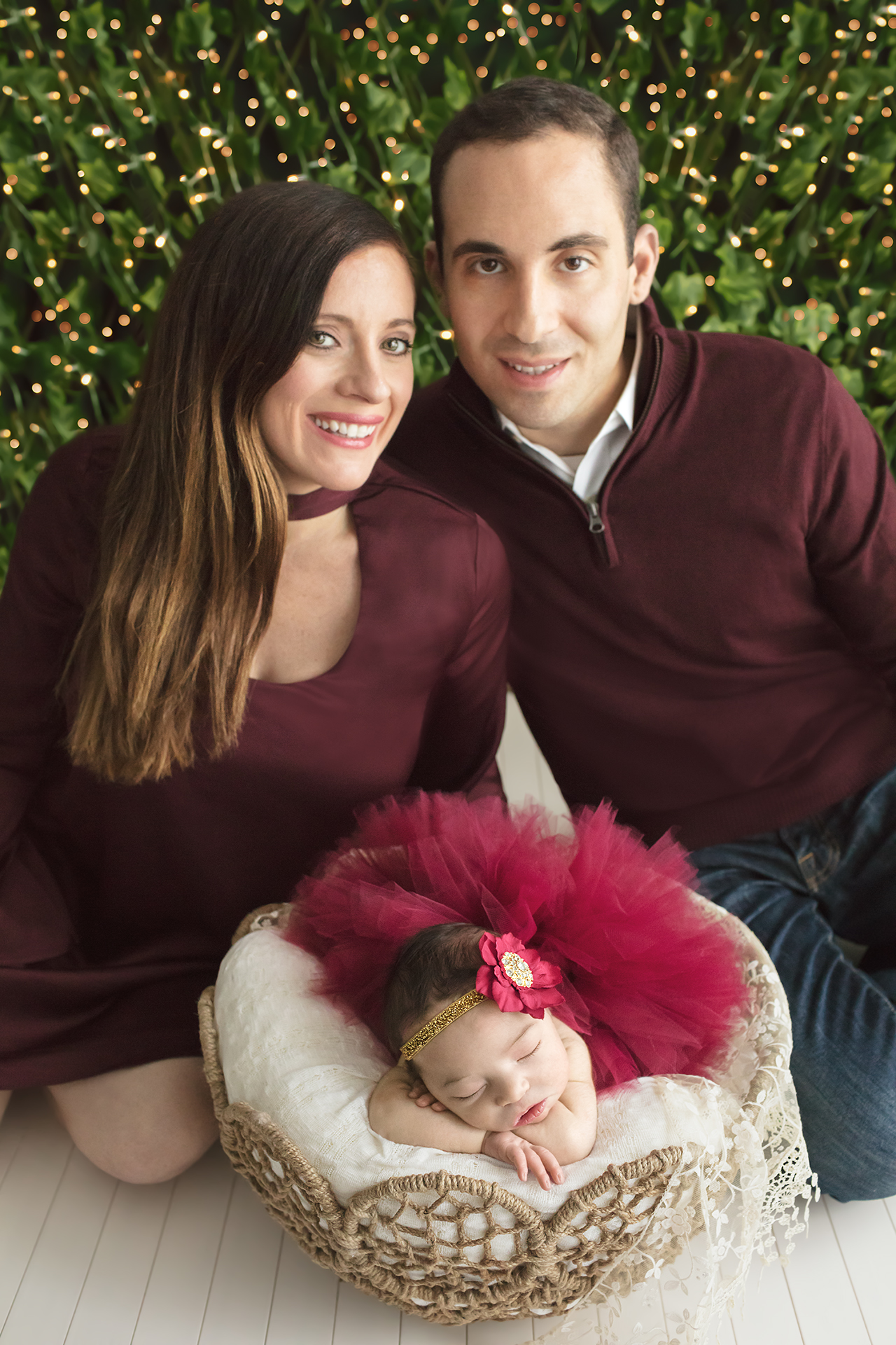 Proud mom and dad with Baby girl posed with red tutu and red flower headband in gorgeous basket in front of twinkle lights for holiday themed newborn portrait session