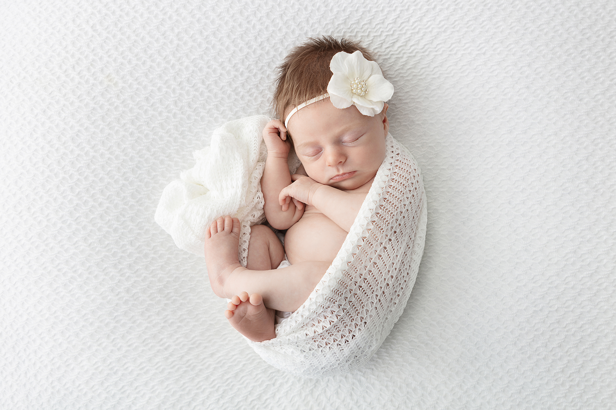 sleeping newborn with a head full of red hair wrapped in an open knit ivory blanket on a woven white background, baby wearing a big white flower headband with pearl flower center