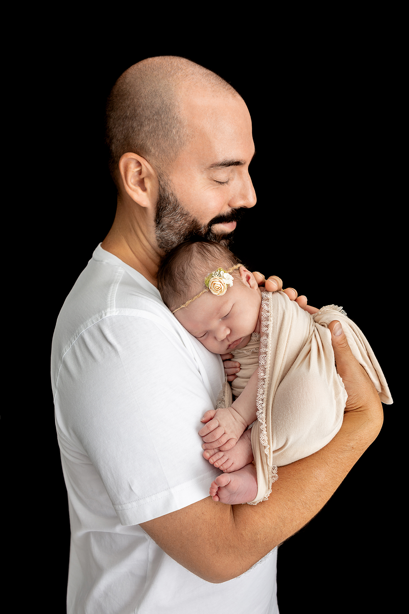 father with a salt and pepper beard wearing a white tee closing his eyes and smiling as he holds his newborn daughter who is wearing a twine and rosette headband and wrapped in an off-white stretch wrap with lace trim, the image is contrasted by a black background