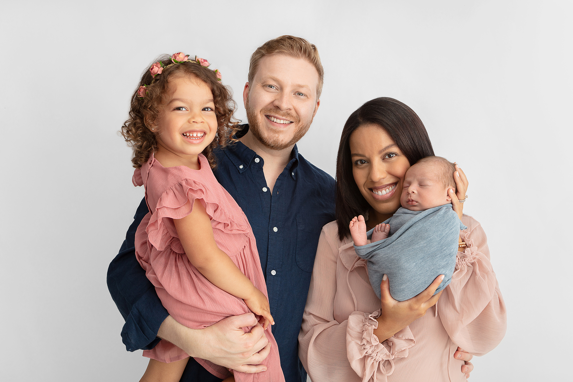 beautiful multiracial family of four smiling at the camera, mom is holding up their newborn who is wrapped in a dusty blue swaddle, soft blue and pink family color palette for photos