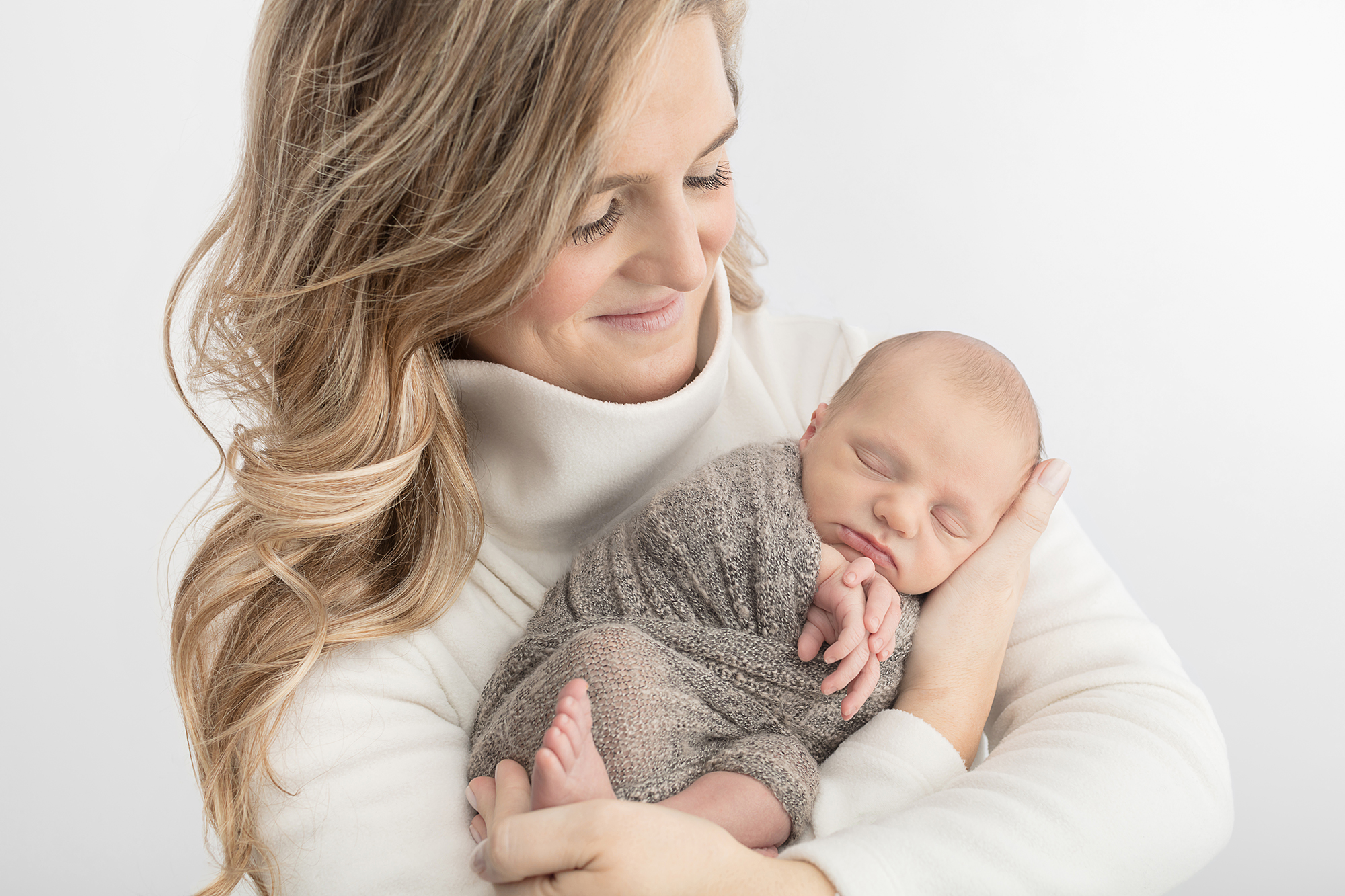 smiling mother with long dimensional blonde hair cradling her newborn who is wrapped in a loose knit gray blanket