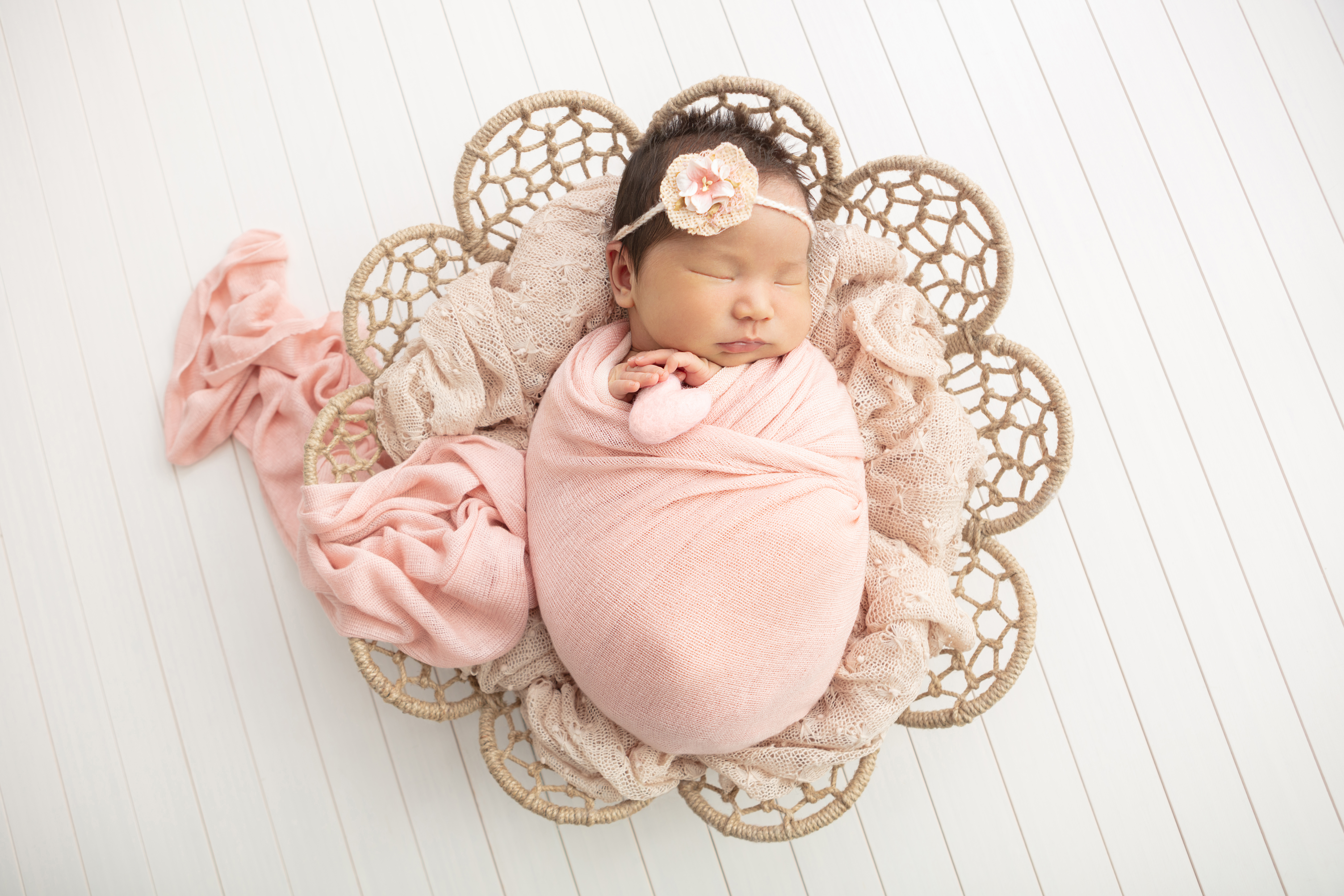 baby girl with dark hair wrapped in a peachy pink swaddle trailing out of a flower shaped wicker basket, rustic white wood floor, delicate peachy pink newborn headband