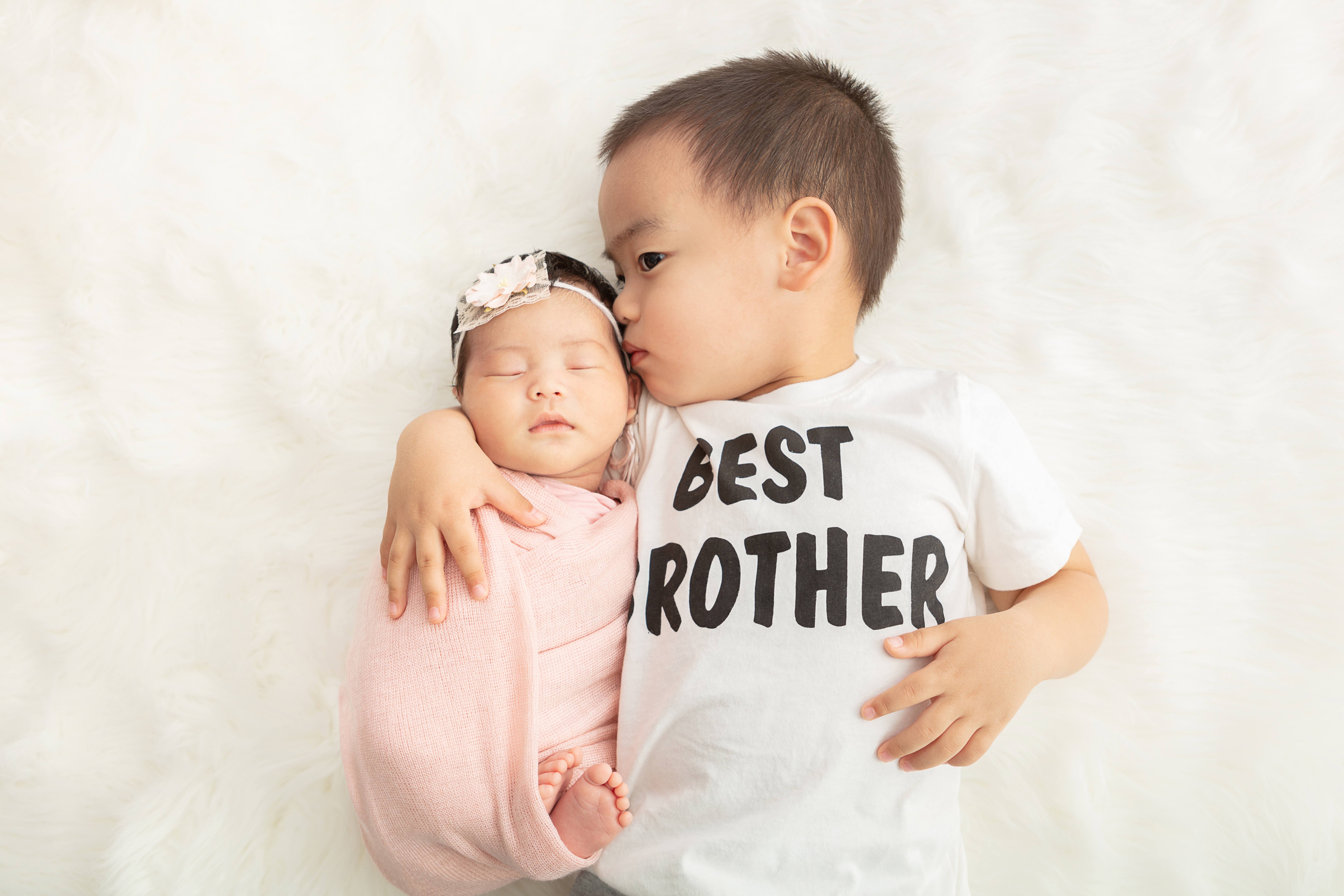 toddler boy wearing a best brother t-shirt and snuggling his newborn baby sister who is wrapped in a peachy pink swaddle