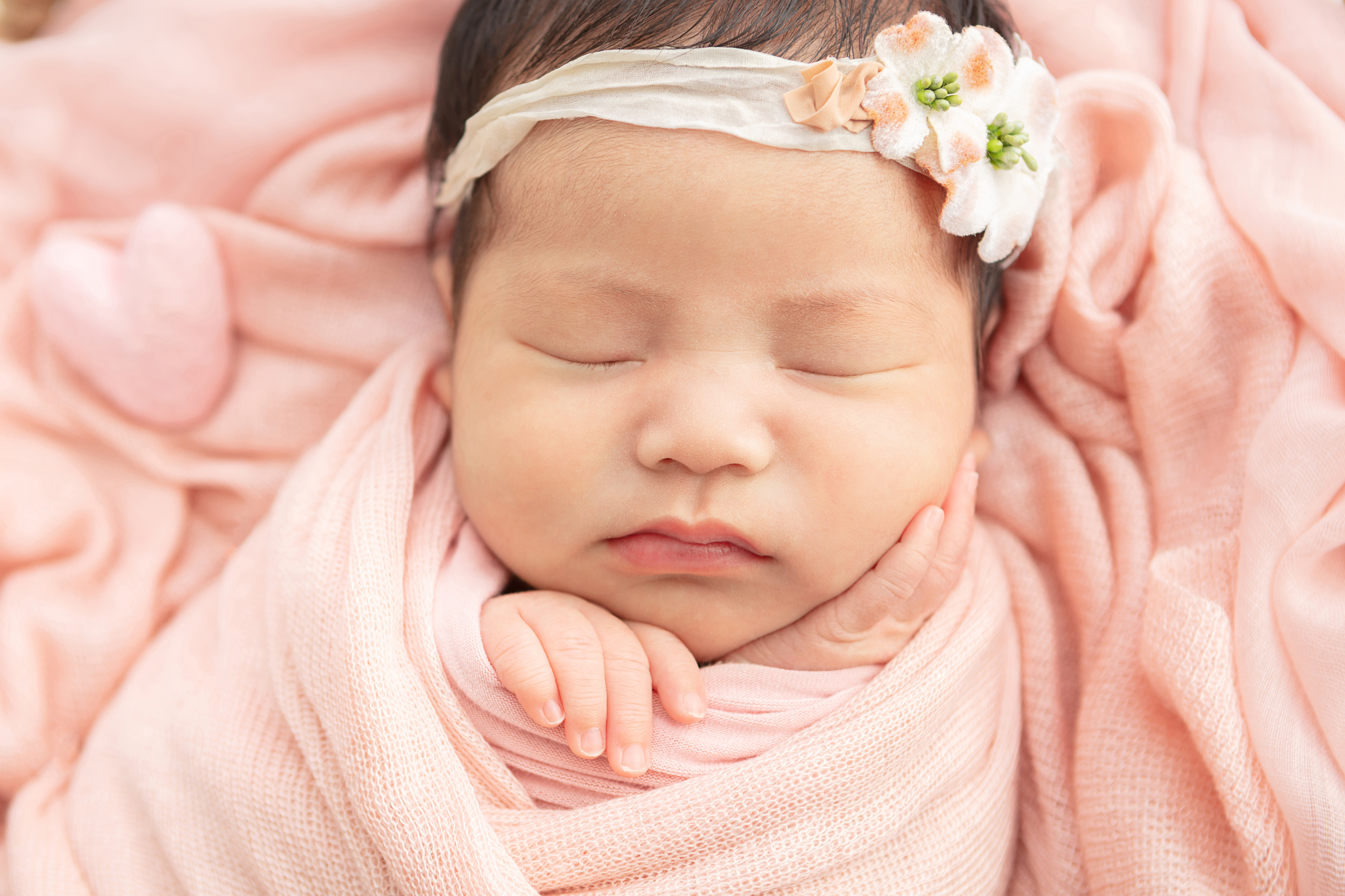 sweet newborn baby girl wearing a muslin floral headband, with her hands delicately tucked in a peachy pink swaddle blanket