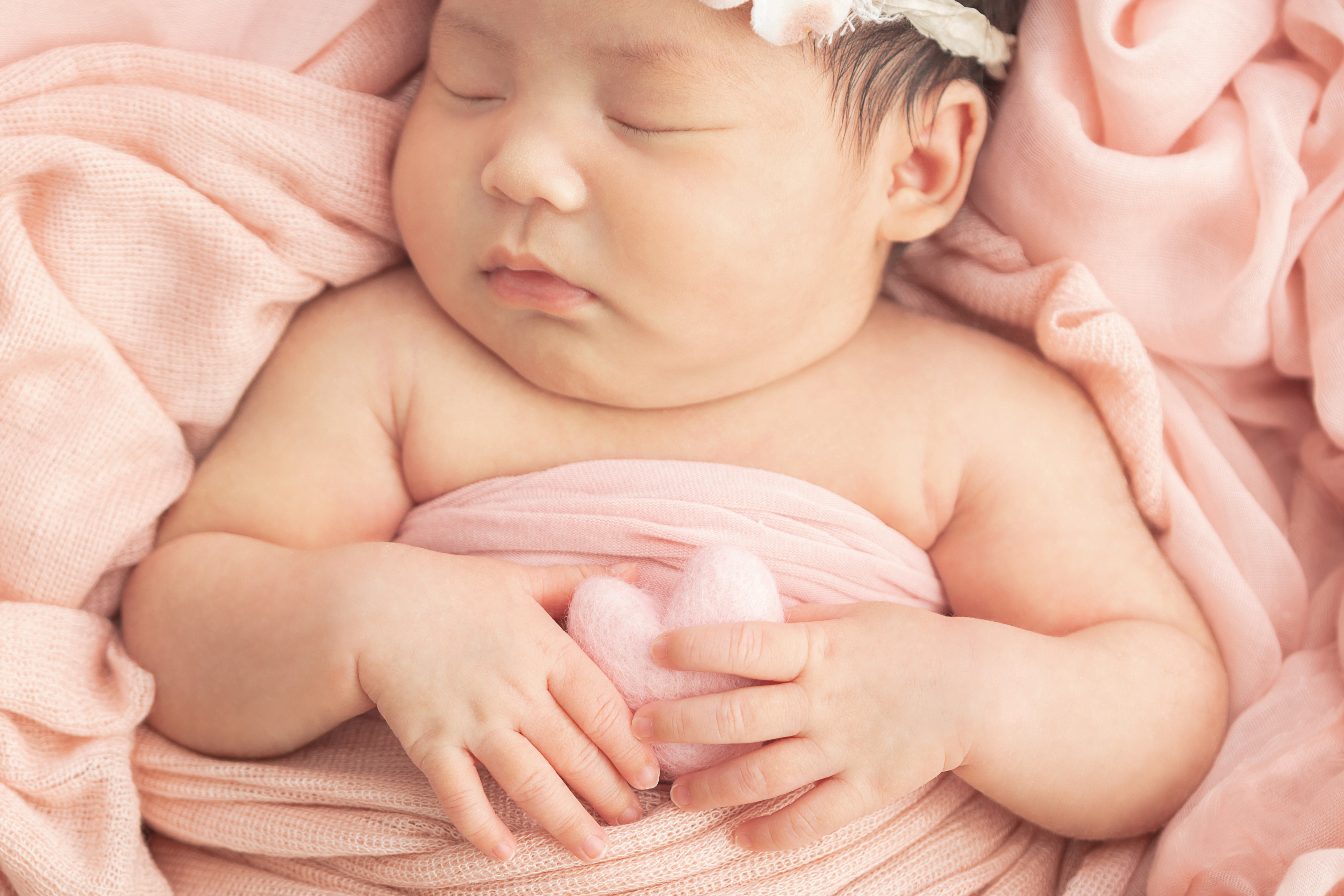chunky newborn baby with her hands gently holding a felted peachy pink heart, napping on a peachy pink swaddle wrap