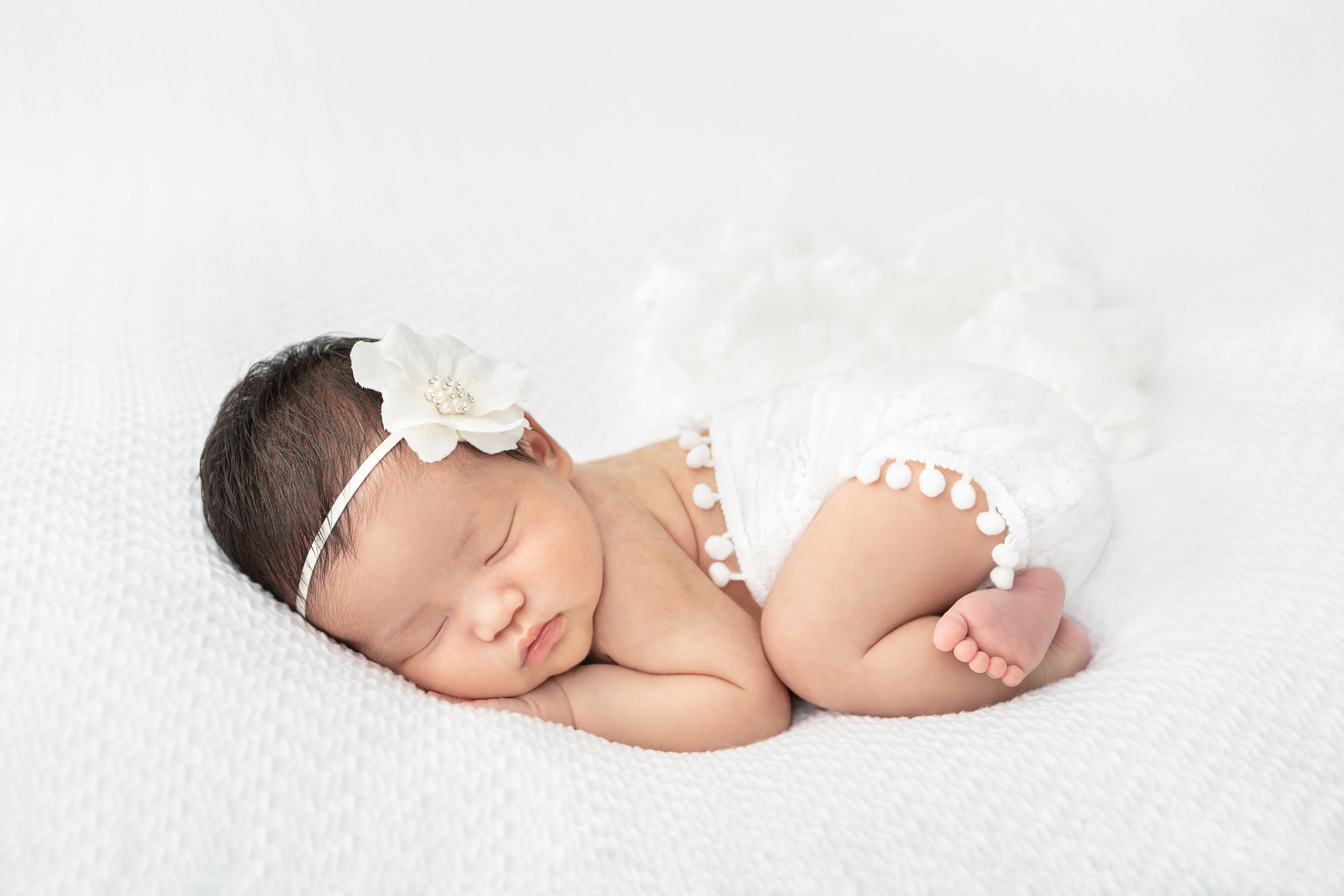 sweet newborn baby girl wrapped in a white swaddle with pom poms and lying on a white textured blanket