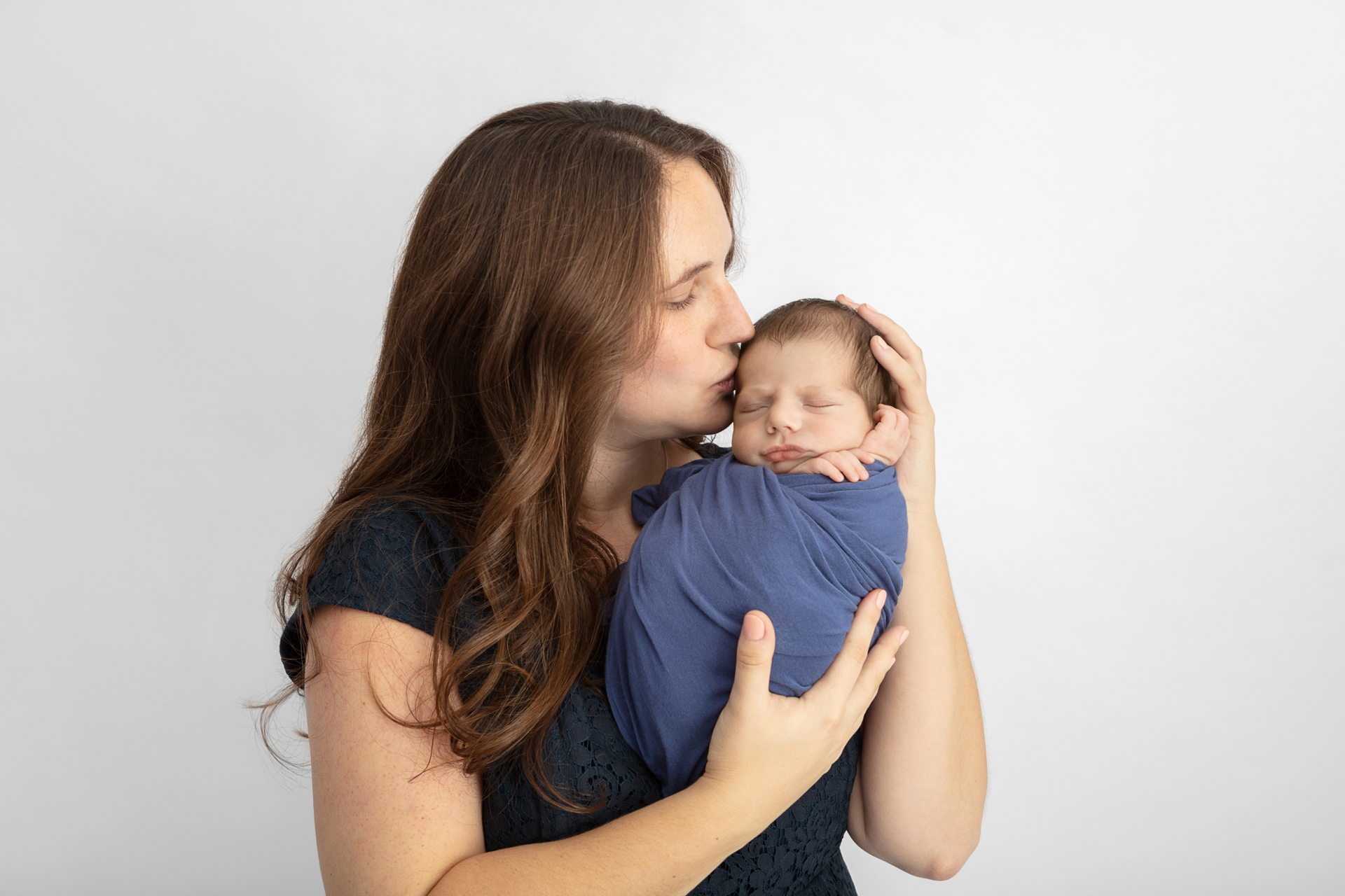 mom with long auburn hair wearing a navy top holding her newborn baby kissing them