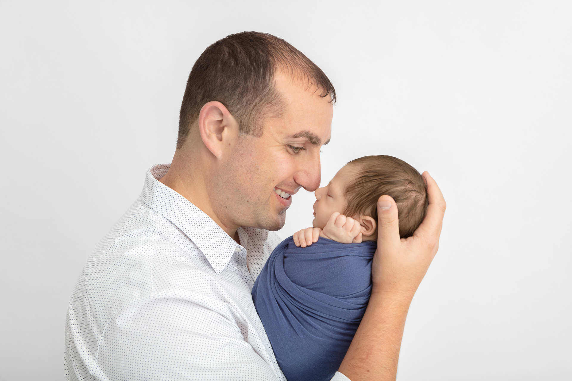 smiling dad holding his newborn son and touching noses with him; neutral studio backdrop