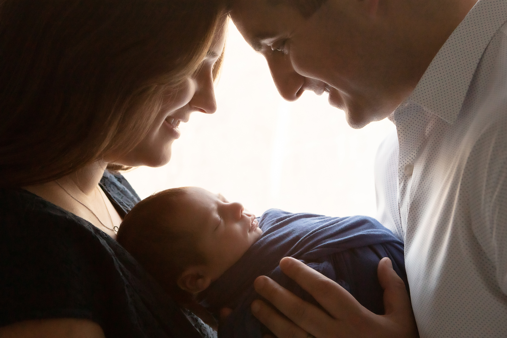 backlit silhouetted photograph of a mother and father smiling and looking down at their newborn son