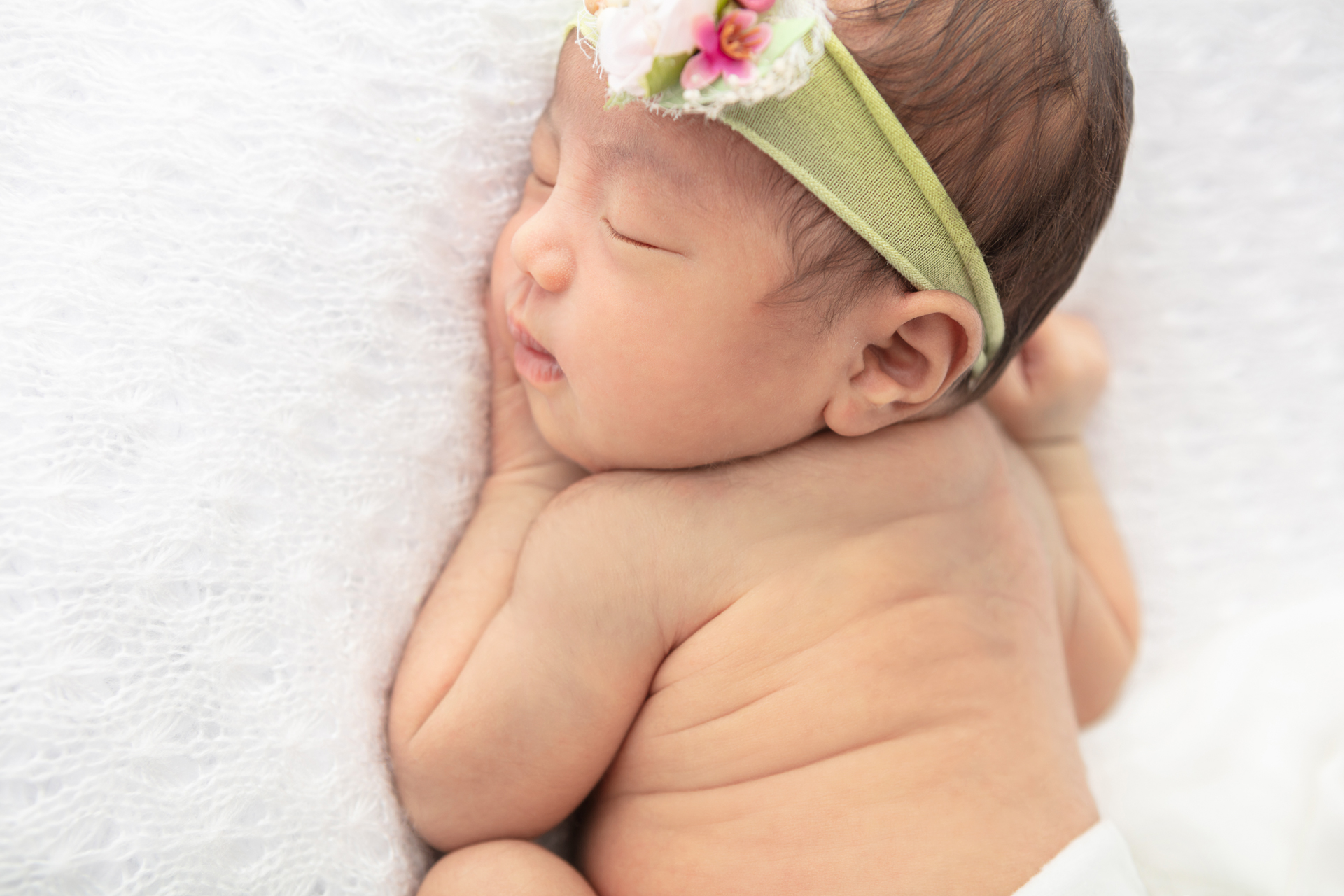 newborn baby girl snuggled up against an open kit white crochet blanket; she is wearing a lime green stretch headband with white and pink faux florals