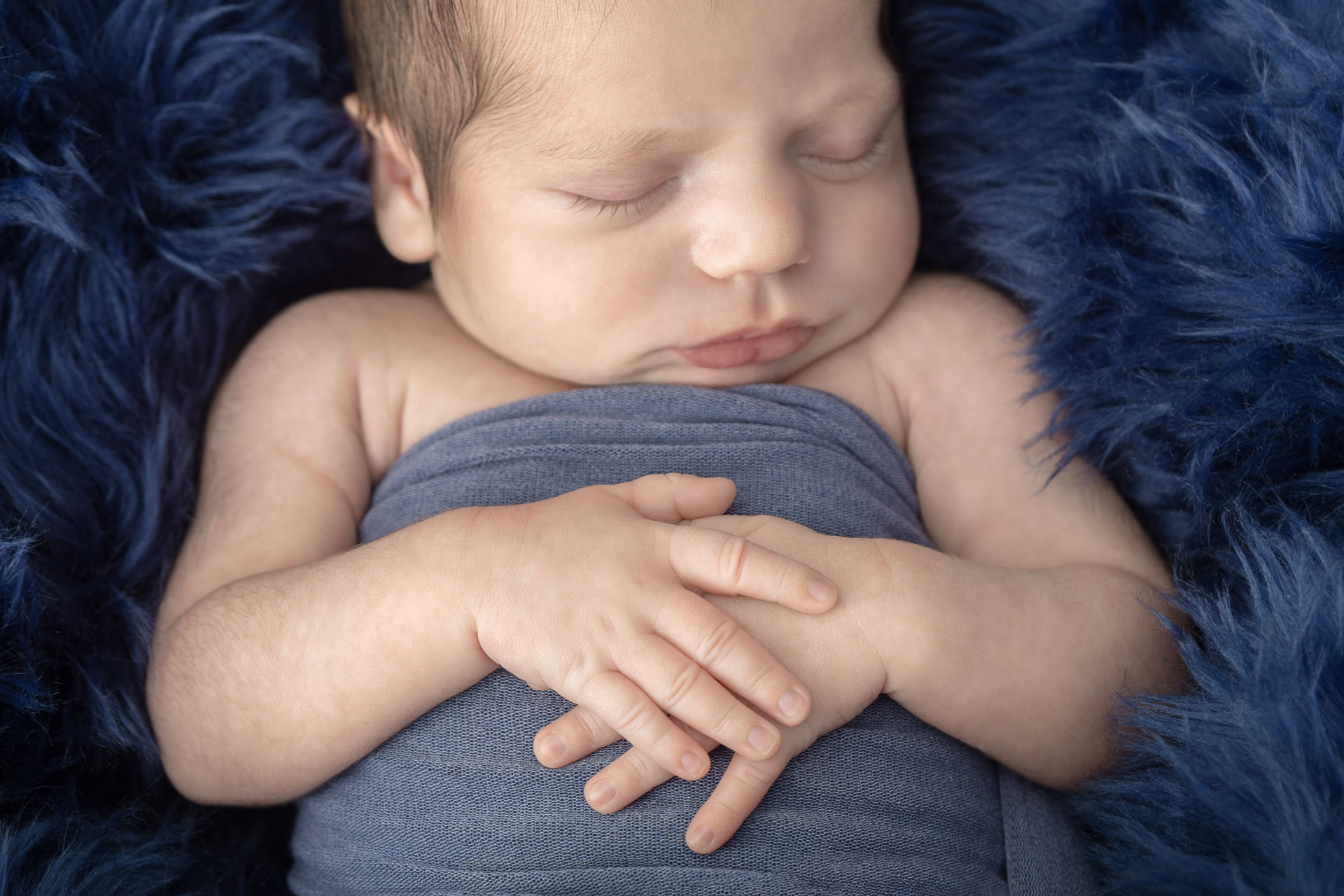 newborn baby boy swaddled in dusty blue with his hands folded across his belly; navy flokati
