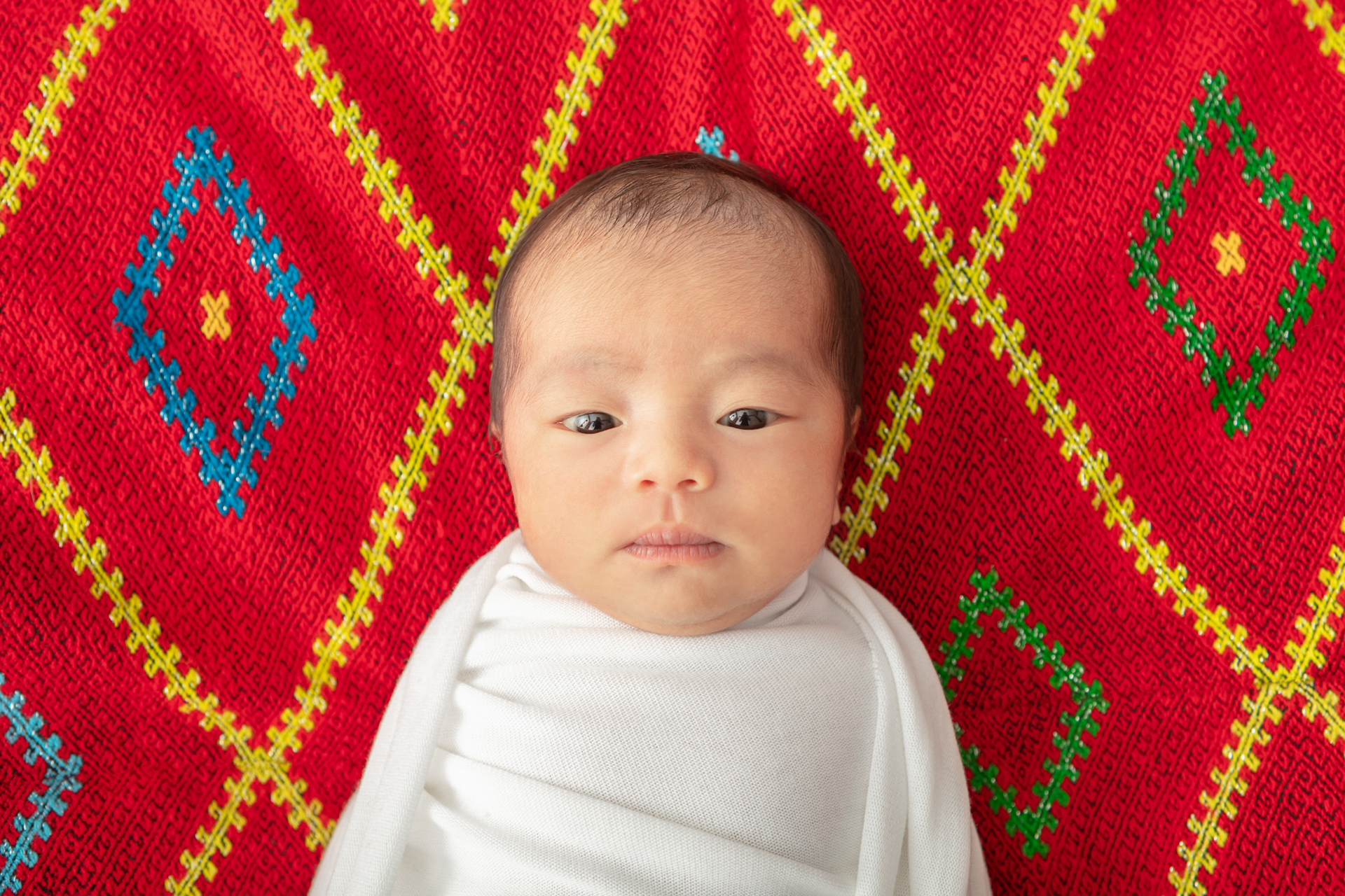 newborn baby girl looking at the camera; she is wrapped in a smooth white swaddle and has a traditional Asian garment behind her; red and blue and yellow and green diamond pattern