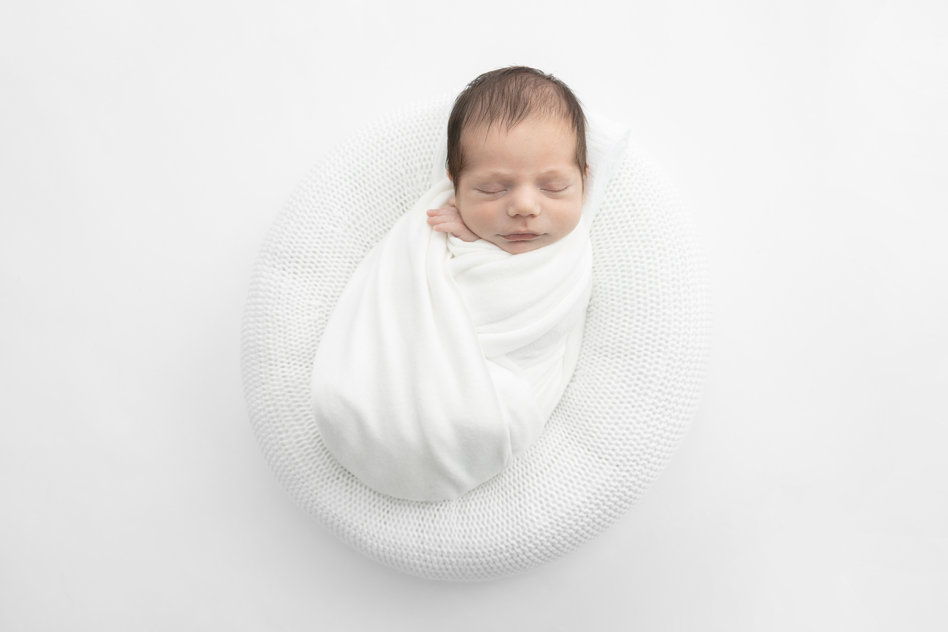 newborn baby wrapped in white asleep on a white knit pouf; white background