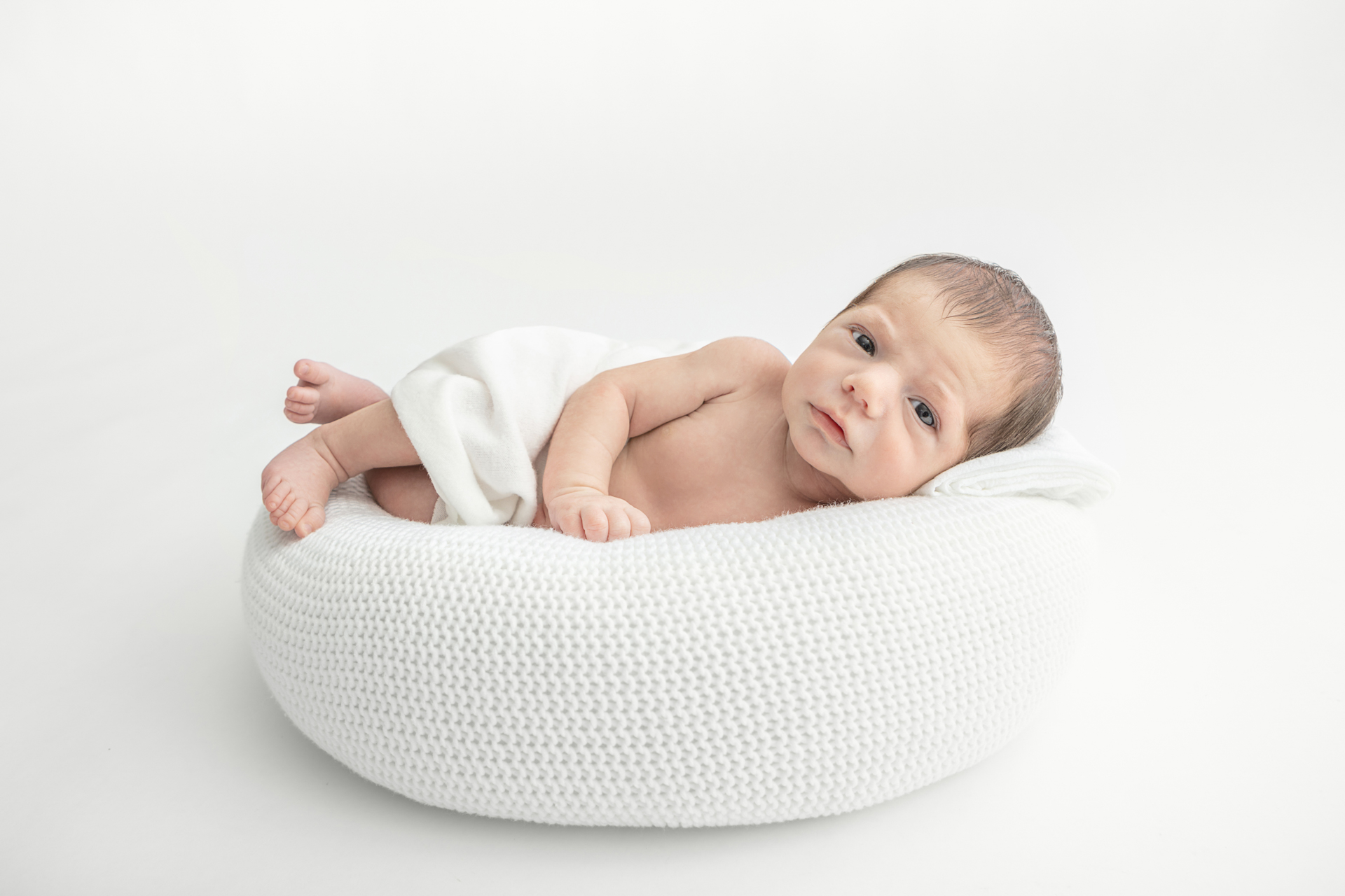 newborn baby looking at the camera with a serious expression; white swaddle; white pouf