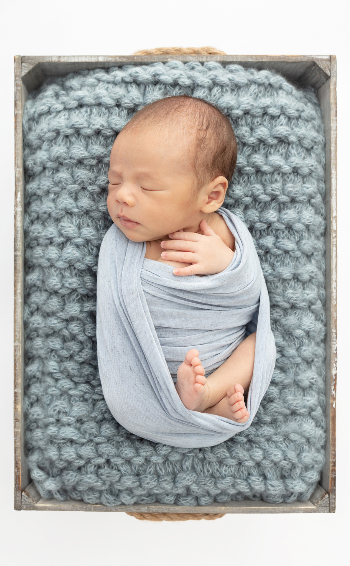 baby boy wrapped in dusty blue, asleep on a chunky knit blanket folded in a rustic box