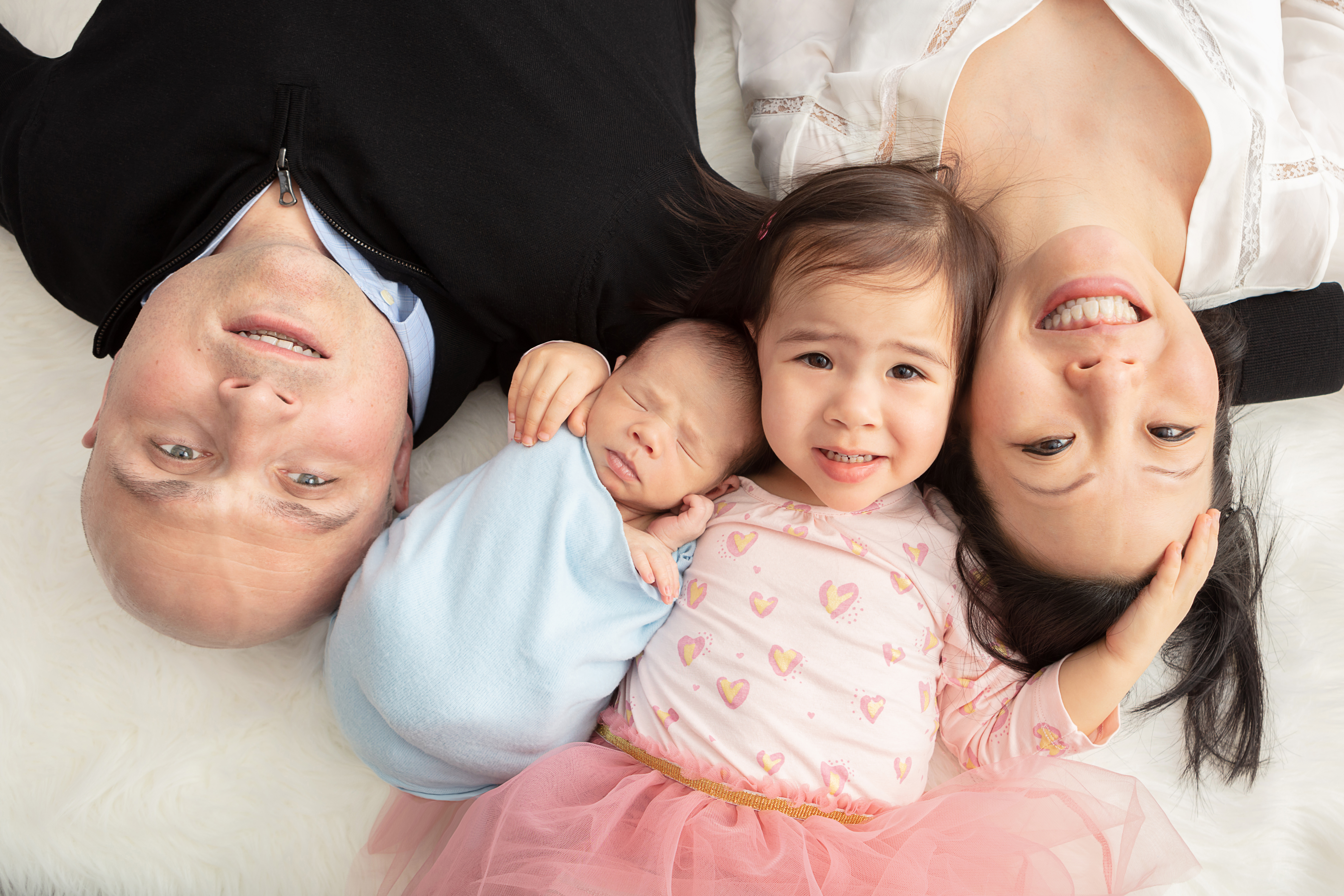 dad, newborn baby boy, big sister, and mom lying on a cream colored blanket and smiling upward