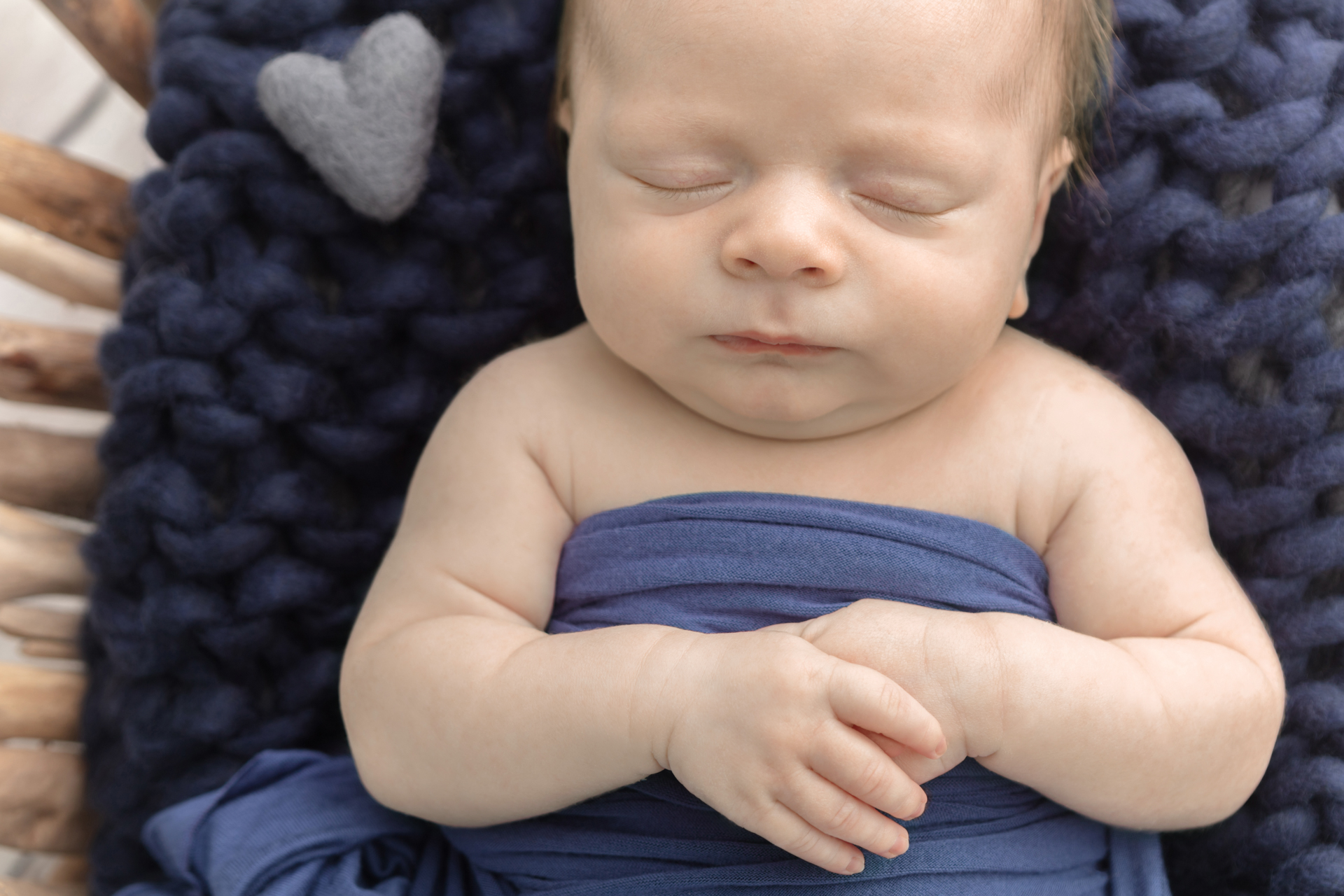 newborn boy with his hands folded across his body, asleep in a driftwood basket lined with a navy chunky knit blanket