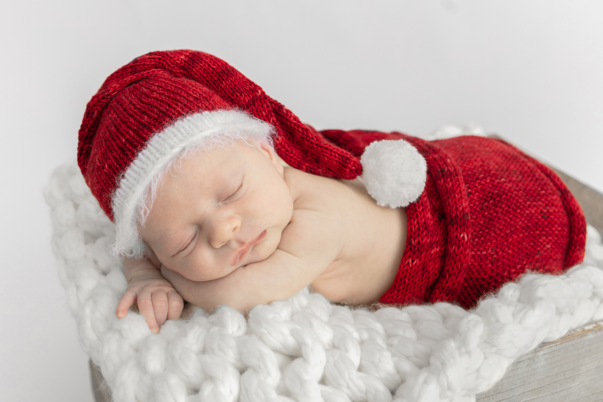 newborn baby asleep in a rustic gray farm box lined with a chunky knit white blanket; newborn boy wearing a chenille Santa outfit