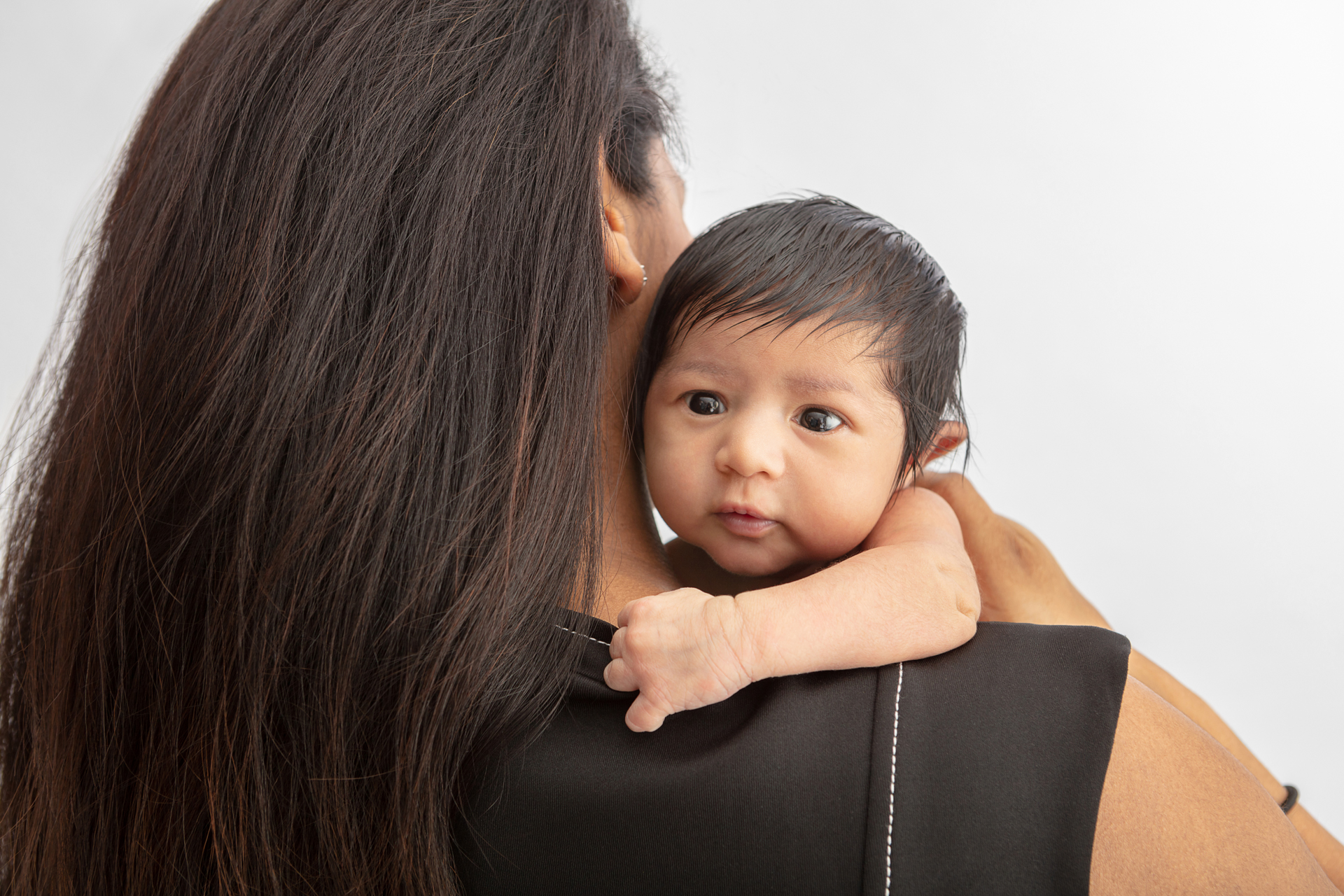 newborn baby girl with dark hair and dark eyes looking over her mother's shoulder