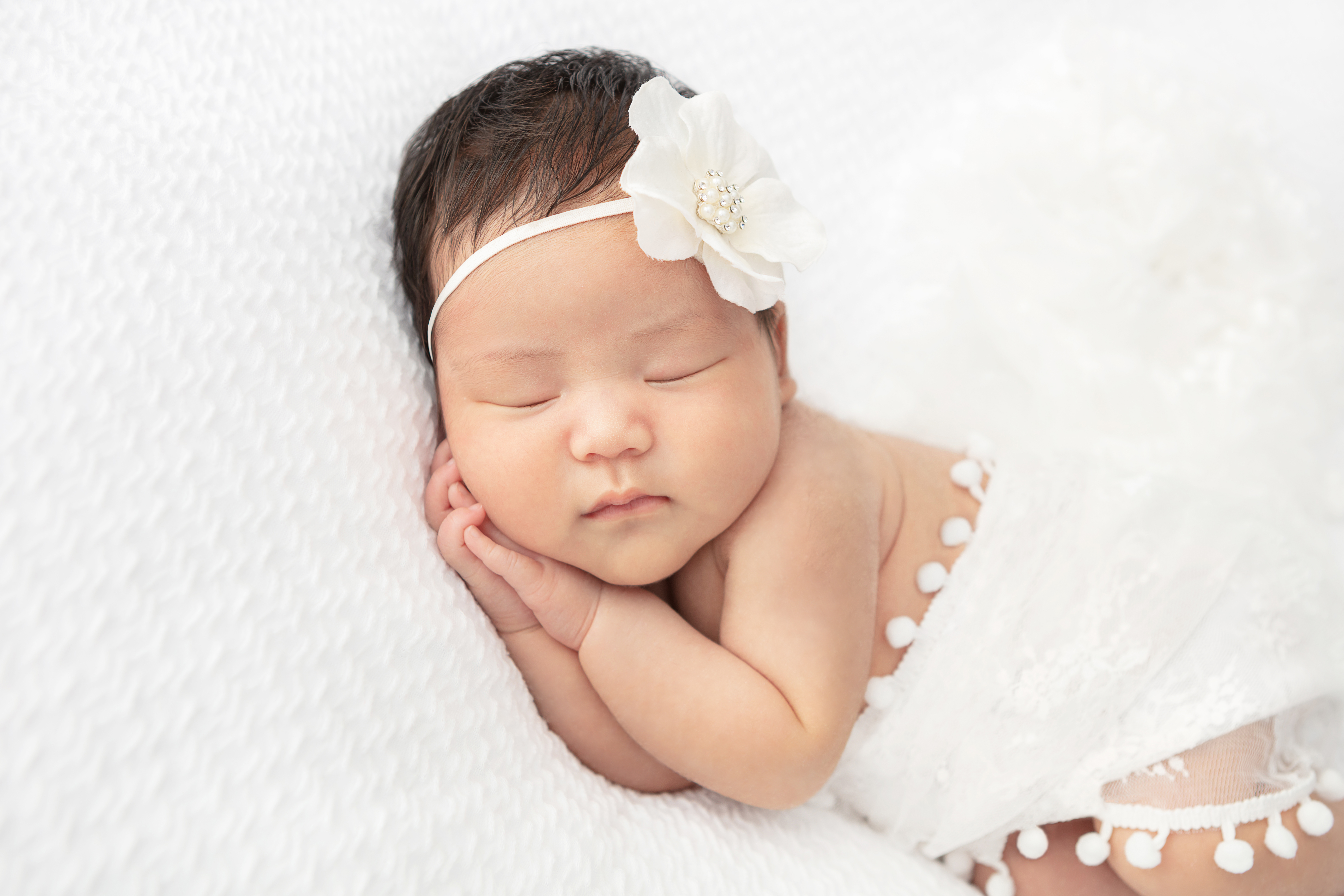 newborn baby girl with dark hair wrapped in a white lace fabric cradled on a soft white fabric withy a white flower headband, hands clasped under her cheek in a sleepy pose