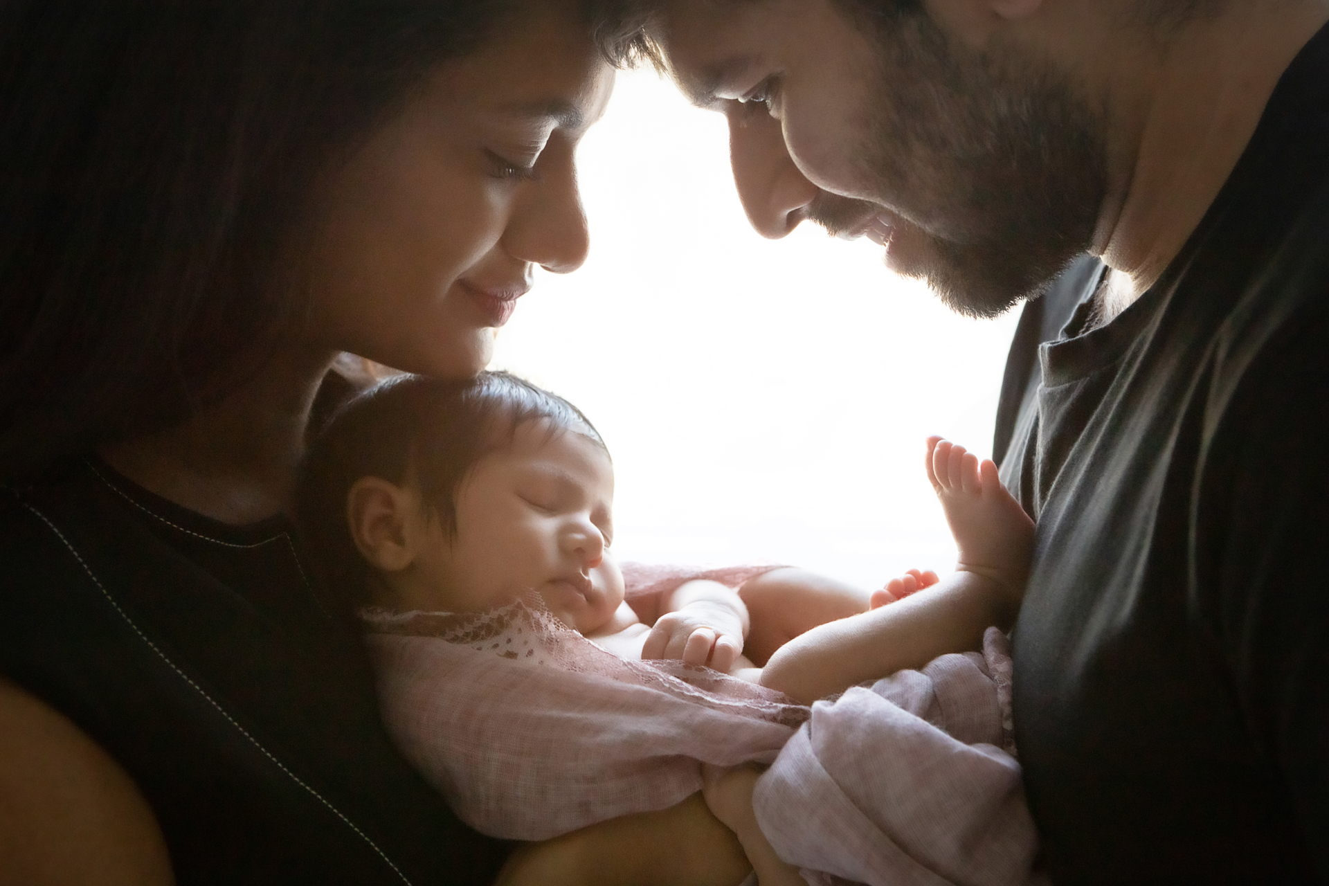 silhouetted image of new parents looking down at their newborn baby girl who is asleep with one foot propped on dad