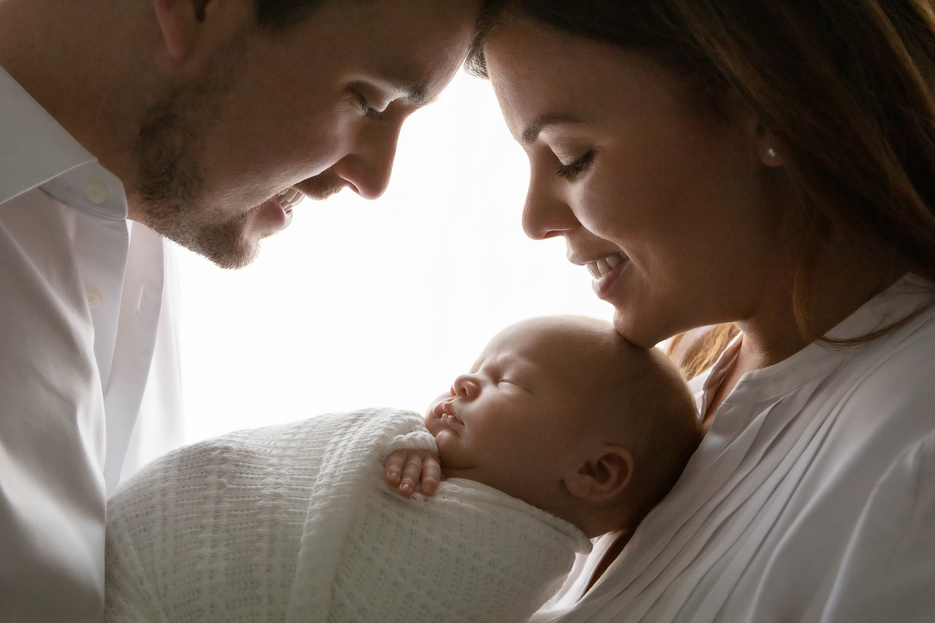 silhouetted, backlit family newborn image; dad, mom, and baby boy asleep with his hand peeking out from the swaddle