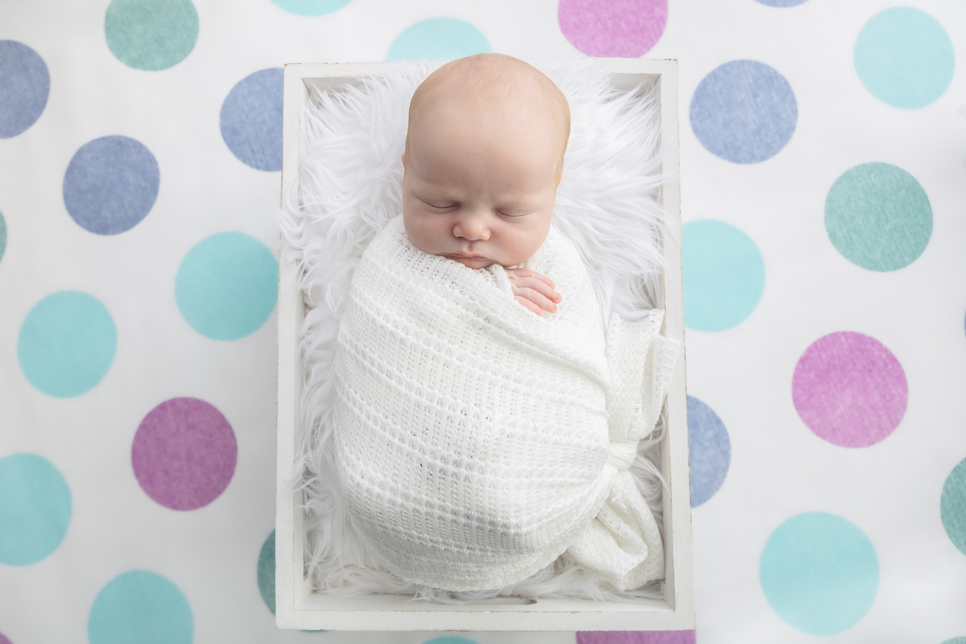 blue, aqua, soft green, and soft purple polka dot background; newborn baby wrapped in an open knit white swaddle
