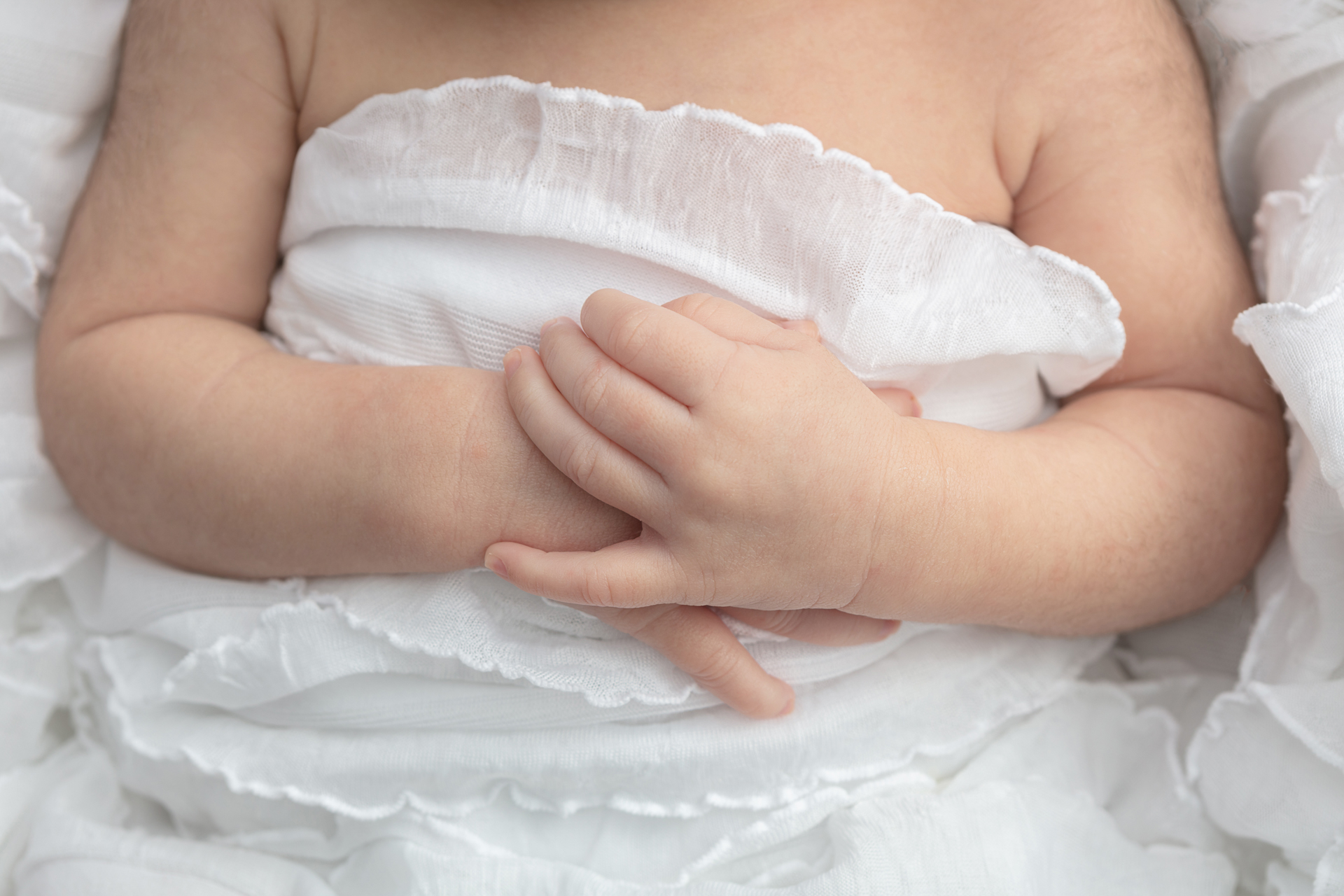 newborn baby hands folded across her belly; white blanket with sheer ruffle details
