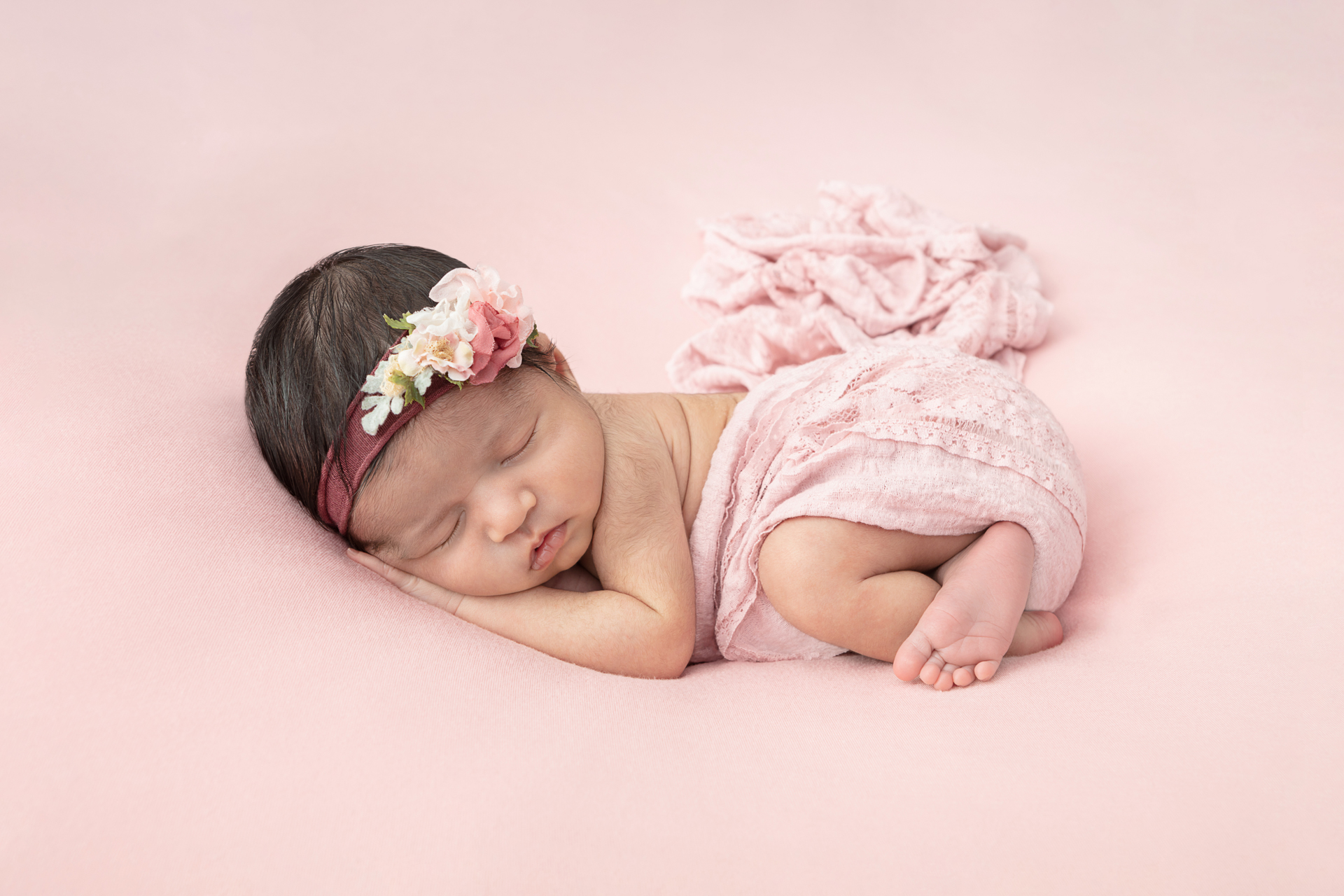 baby girl asleep on a pink backdrop and wrapped in a pink swaddle while wearing a floral headband
