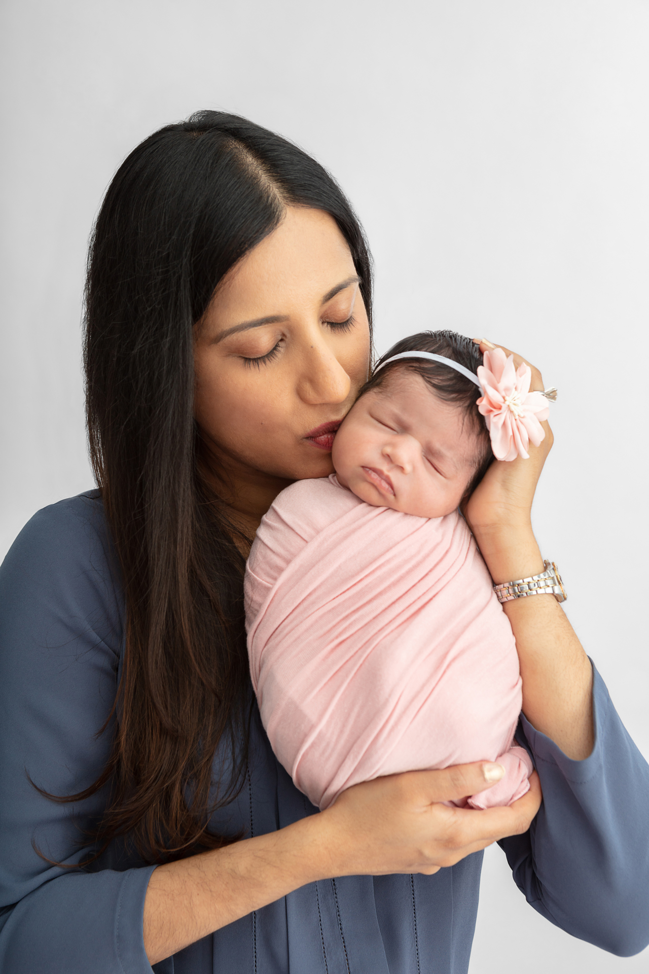 new mom with long dark hair holding up her newborn baby girl who is swaddled in light pink