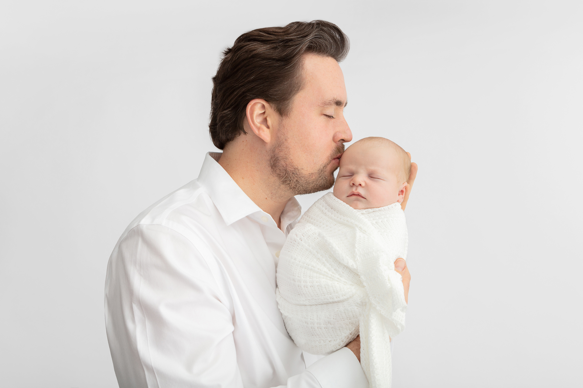 new dad in white shirt holding his son up and kissing his head; baby boy wrapped in white