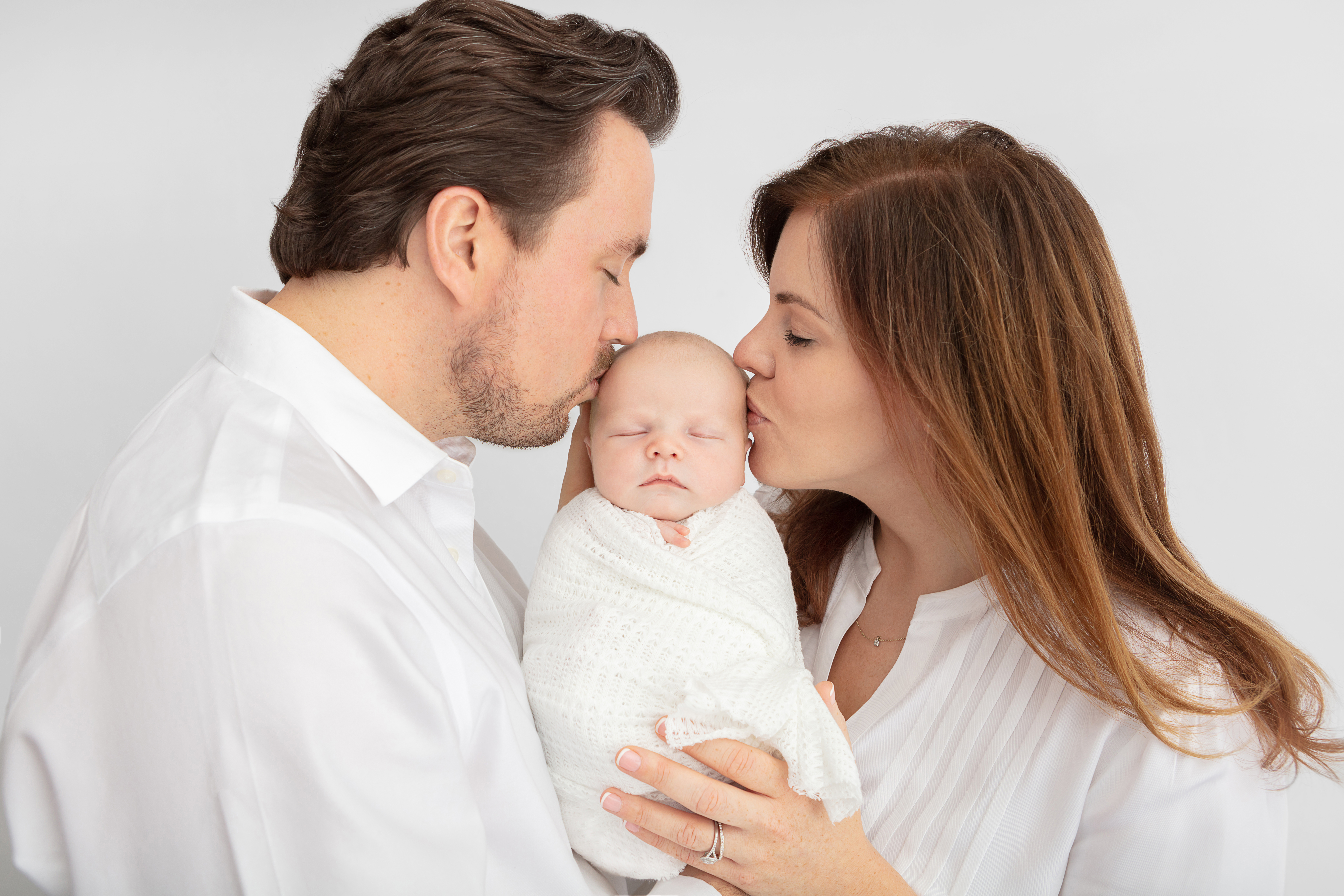 Studio posed newborn baby portrait session all in white, baby is cradled in-between mom and dad, kissing him on the cheeks
