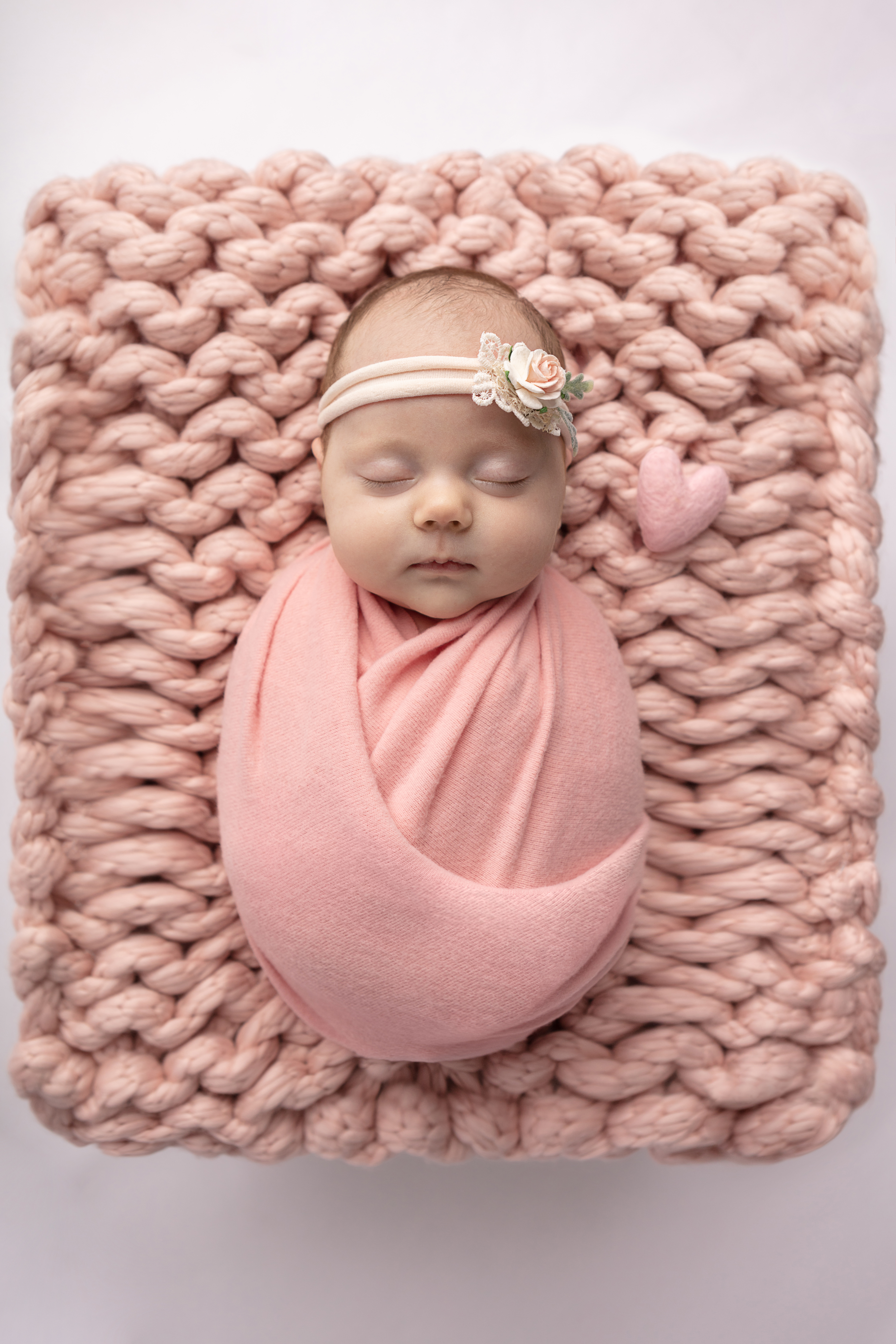 newborn baby girl swaddled in dusty pink, wearing a stretch floral headband; pink chunky knit blanket