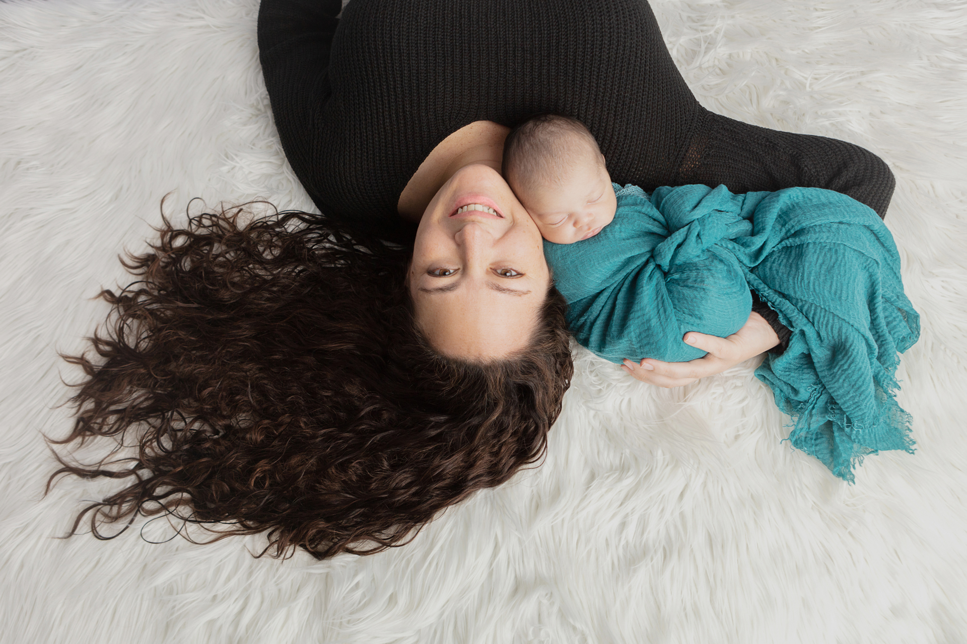 simple and classic portrait of a new mother with long and wavy mermaid hair holding her new baby boy while laying on a white flokati rug