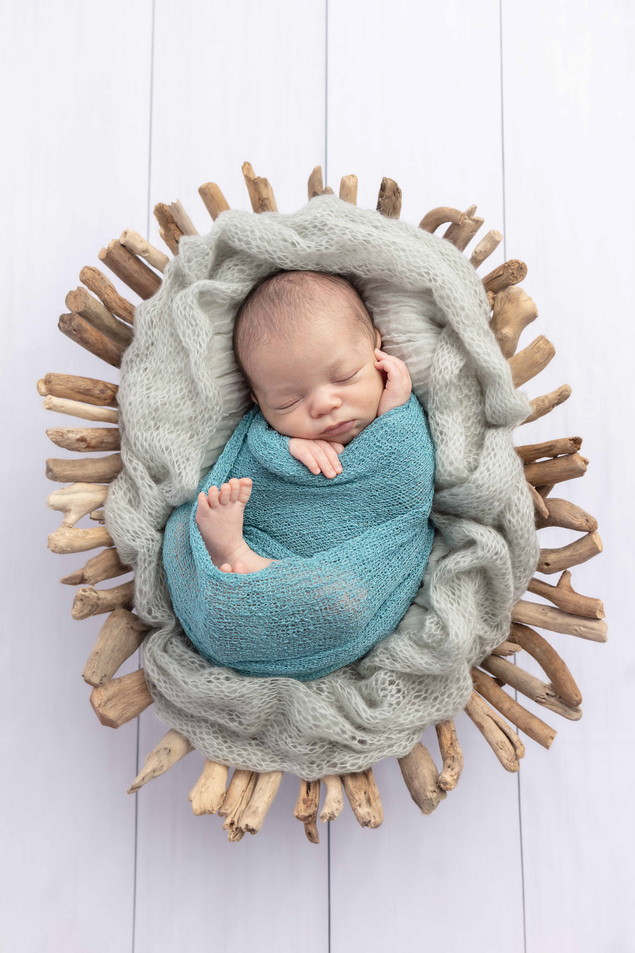 baby boy swaddled in an open knit blue swaddle, in a driftwood basket on a white wood floor; Looking Up Photography