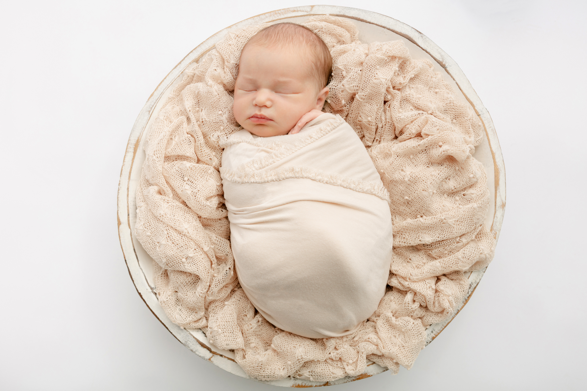 newborn baby girl swaddled in a neutral lace-trim swaddle, asleep in a large basin bowl