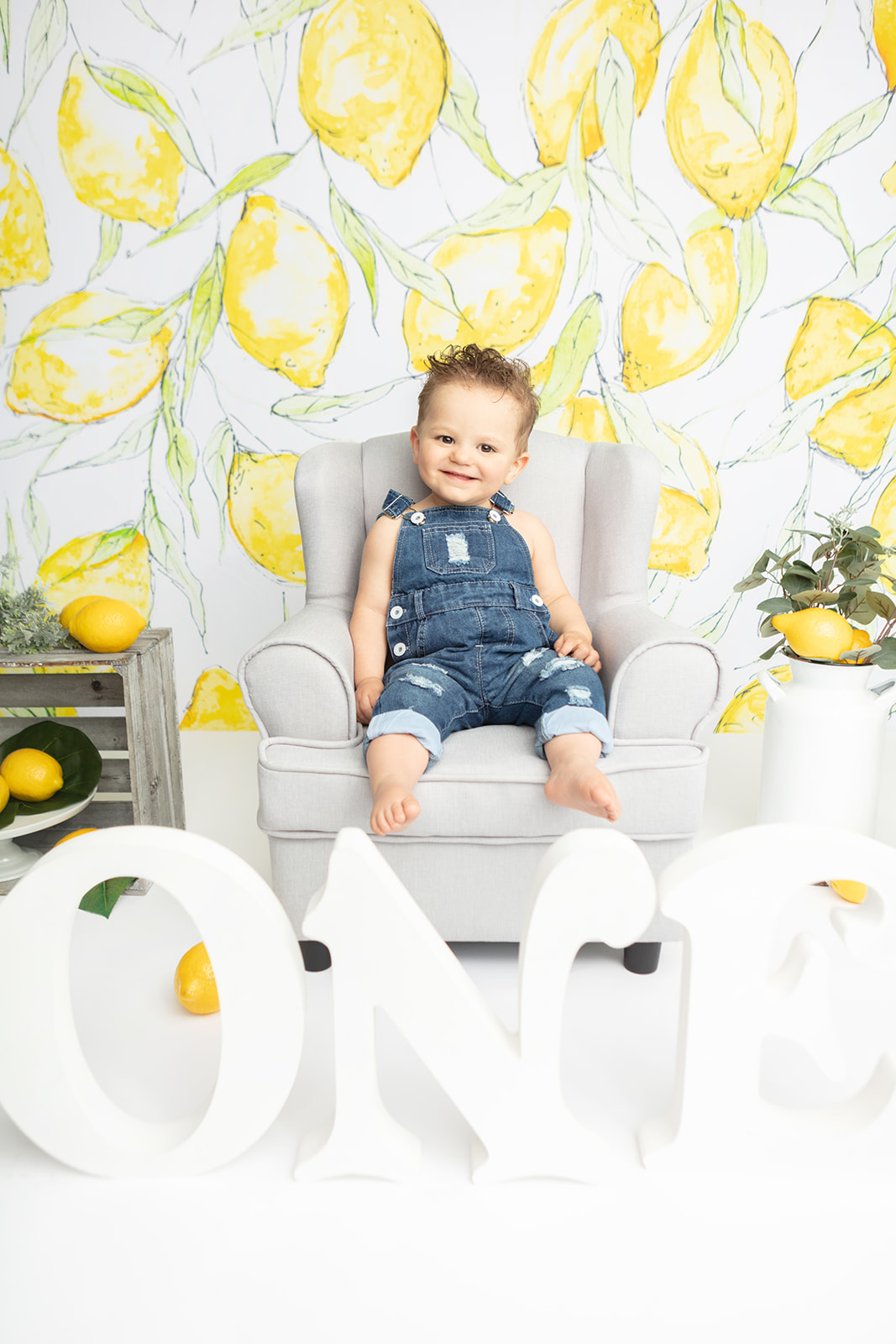 A smiling little boy in denim overalls sits in a light gray easy chair. In the foreground of the image are oversized, styled letters spelling out the number "one". A lemon backdrop by Heidi Hope and farm style lemon accessories add visual interest to the photo.