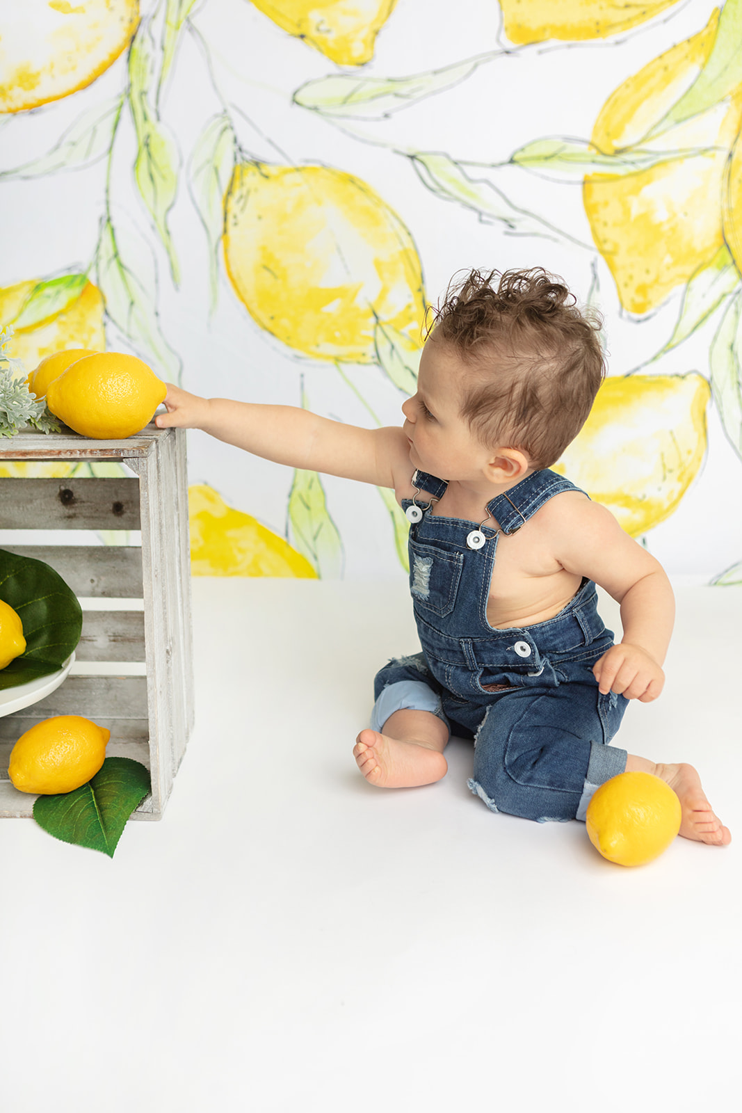 A sitting, one year-old little boy wearing denim overalls, reaches for a lemon prop during his lemon themed first birthday cake smash session with Karen Kahn, of Looking Up Photography.